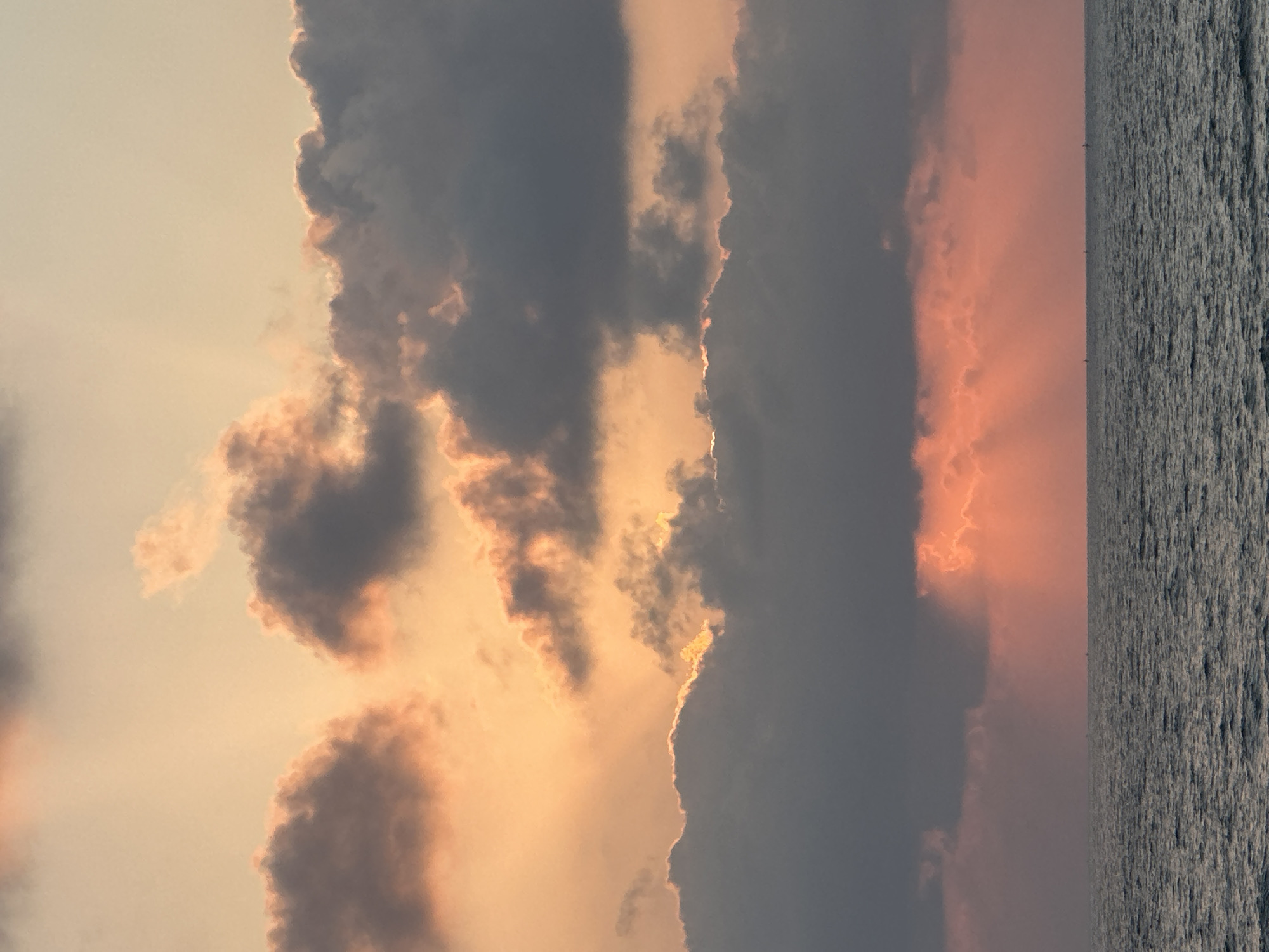 The image depicts a dramatic sunset over the sea, likely in the Mate Asher Regional Council, Israel, given the context. The sky is filled with a vibrant array of colors, from soft oranges and pinks to deep grays and purples in the clouds. Sunlight pierces through the cloud cover in distinct rays, creating a luminous effect. The sea below is a deep, textured gray, with small whitecaps visible. On the horizon, several distant, dark silhouettes suggest small boats or possibly distant landforms. There are no people or discernible structures visible in the immediate scene.