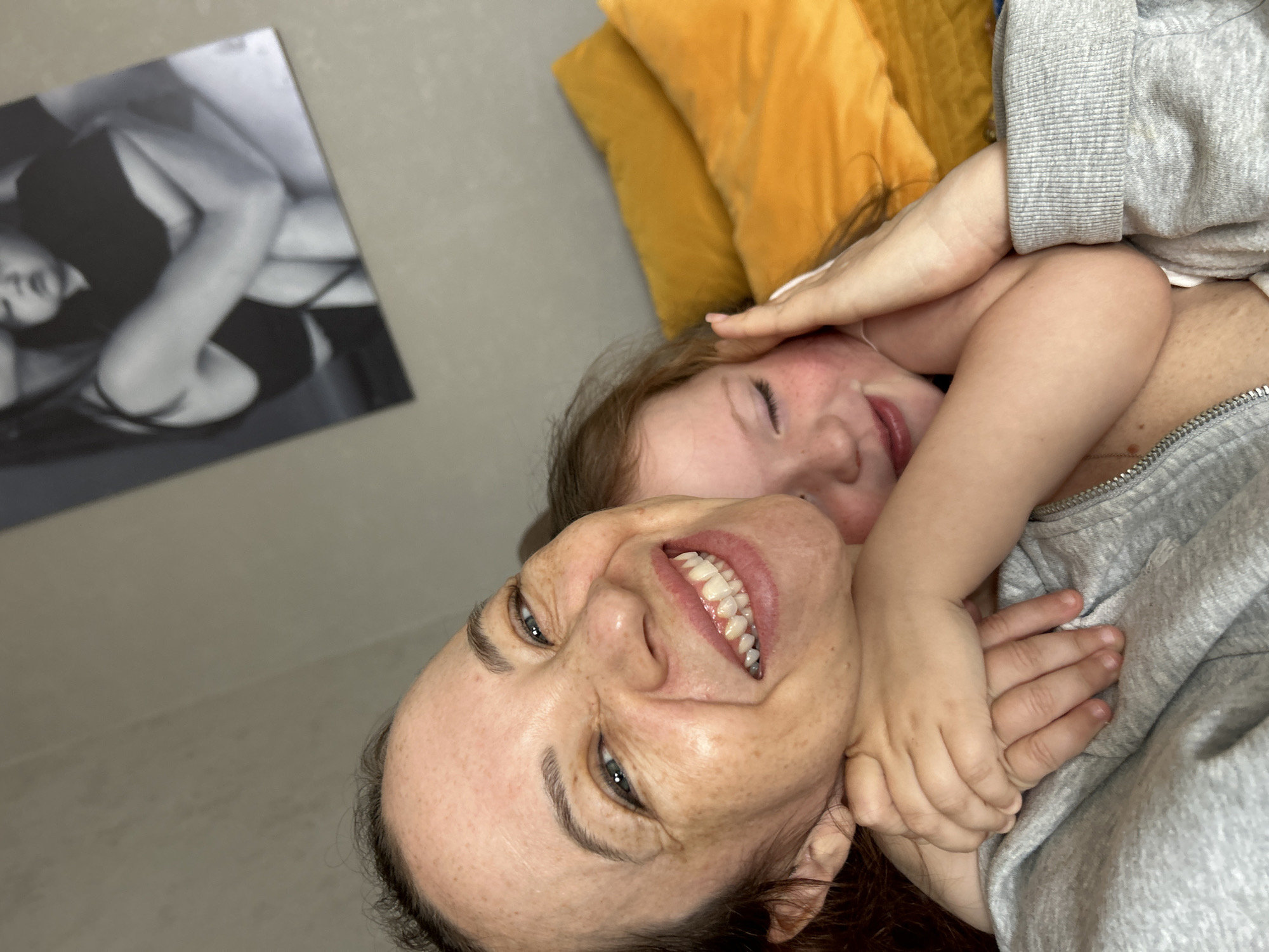 A woman and a child are lying down, seemingly embracing. The woman is facing the camera with a broad smile, revealing her teeth. She has fair skin with visible freckles and wrinkles. Her eyes are open and appear to be grey. The child, with fair skin and reddish-brown hair, lies partially on top of the woman, with arms around her neck. The child's eyes are closed. The woman is wearing a grey zippered jacket. Behind them are yellow pillows. On the wall in the background hangs a black and white poster featuring a woman. The location is Chaikovsky, Russia.