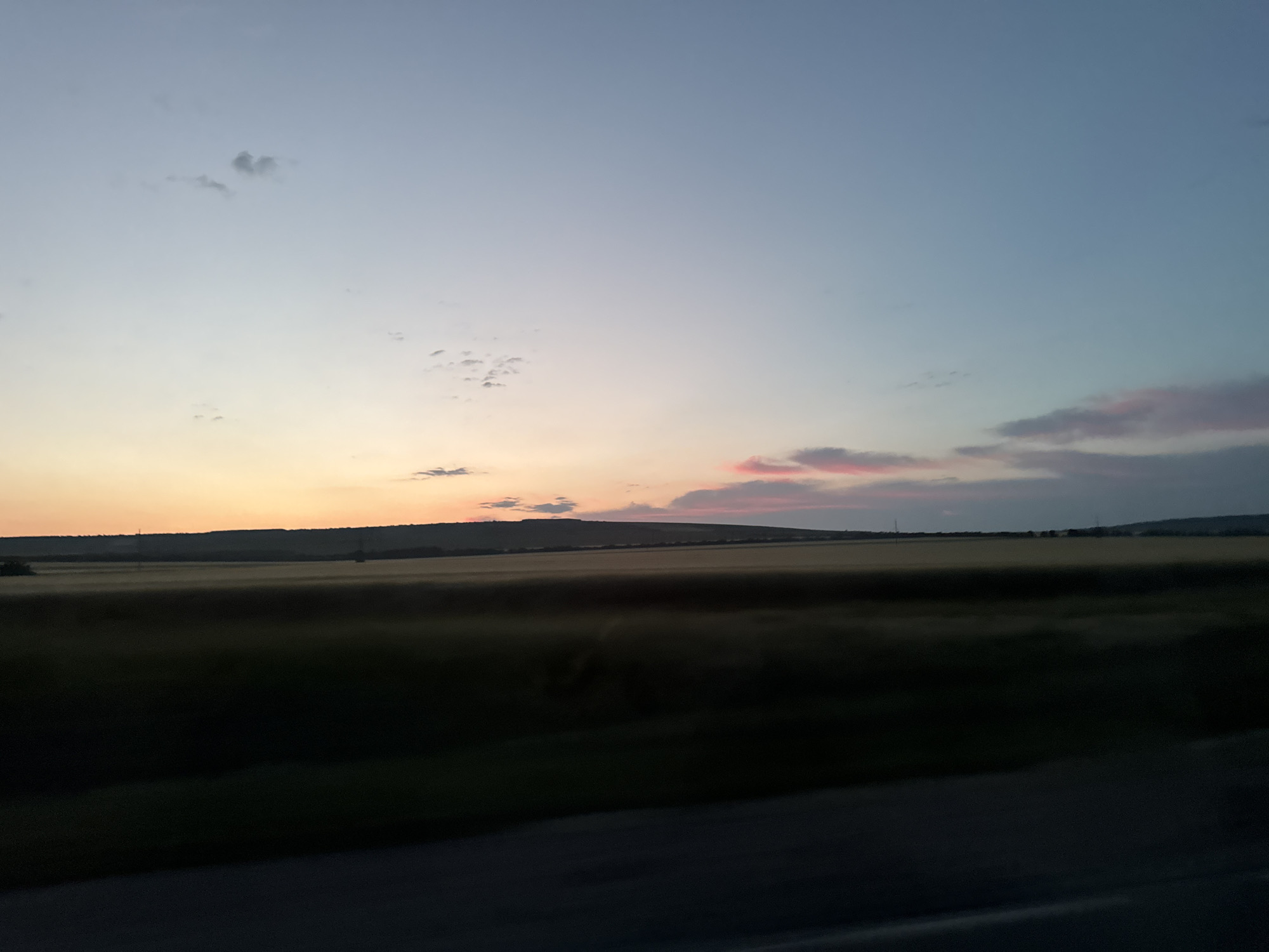 The image depicts a rural landscape at dusk or dawn. The sky transitions from a pale orange at the horizon to a soft blue overhead, with scattered wispy clouds tinged with pink. The foreground is dominated by a dark, indistinct mass, likely foliage or a road, with a blurry, sunlit field stretching into the middle ground. In the distance, a low-lying horizon of hills or trees is visible, silhouetted against the colorful sky. The overall impression is one of a peaceful, natural setting. The location is not specified and is listed as Unknown, Unknown.