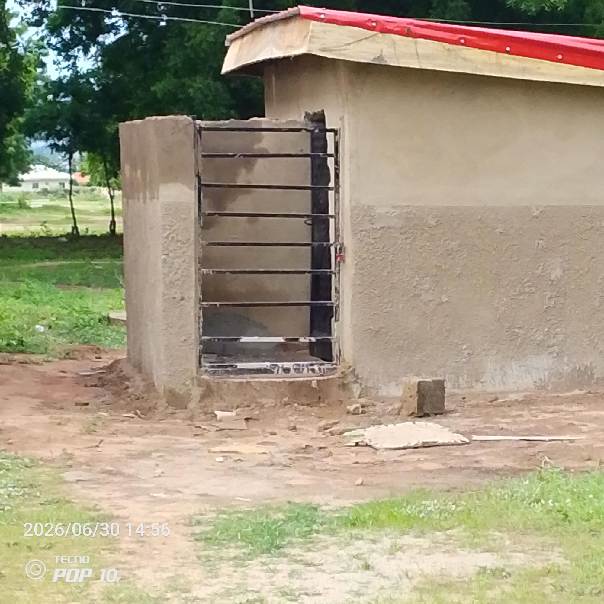 The image depicts a rustic structure with a red-tarped roof, possibly under construction or renovation, situated in an outdoor setting. The building appears to be made of rough-textured, light-colored material, with a metal grate covering an opening on its side. The ground surrounding the structure is dirt and gravel, with patches of green grass and some debris. In the background, trees and some distant buildings can be seen, suggesting a semi-rural or developing area.
The scene appears to be a daytime shot, with ample natural light. The weather seems to be pleasant, given the green vegetation. There are no people visible in the image. The overall impression is one of an undeveloped or transitional area.
Visible text in the image includes a timestamp: "2026/06/30 14:56", and a device indicator: "TECNO POP 10". This text suggests the image was taken by a Tecno Pop 10 mobile phone on June 30, 2026, at 2:56 PM. The location context provided is Jalingo, Nigeria.
