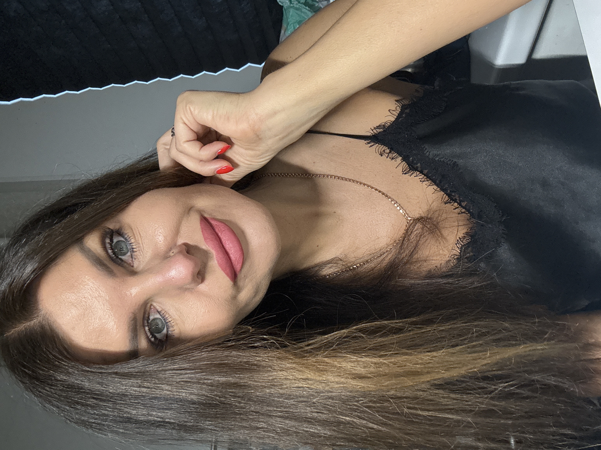 The image shows a close-up of a woman with long, brown hair and blue eyes. She is wearing a black camisole with lace trim and a delicate gold necklace. Her lips are painted a matte pink, and her fingernails are painted red. She is touching her hair and appears to be looking at the camera with a slight smile. The background is dimly lit and appears to be indoors, possibly a room in a home. There are no specific details in the image that indicate the location in Chaikovsky, Russia.