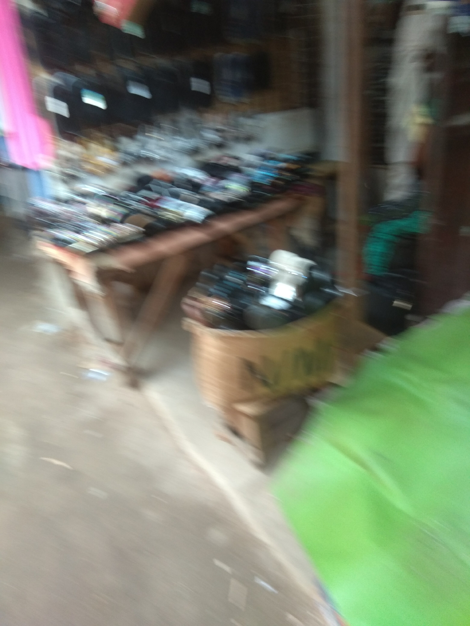 The image depicts a blurry, indoor scene that appears to be a market stall or shop in Jalingo, Nigeria. The primary subjects are various objects, including what seem to be clothing items hanging on display, possibly on racks or shelves, and a collection of items in a basket. There are also some bags and what looks like electronics or accessories.
The lighting suggests it is daytime, and there are no clear indications of weather conditions. The overall impression is of a bustling, albeit somewhat disorganized, retail environment. The blurriness of the image, possibly due to motion or a low-quality camera, makes it difficult to discern specific details about the objects or any people who might be present. However, the presence of clothing and merchandise points to a commercial setting where goods are being sold.
There is no visible text in the image. The emotional tone is neutral, as the blurriness prevents any clear facial expressions or gestures from being observed. The setting appears to be a covered area, typical of market stalls or small shops in warmer climates. The arrangement of items suggests a retail space that might be packed with a variety of goods for sale.