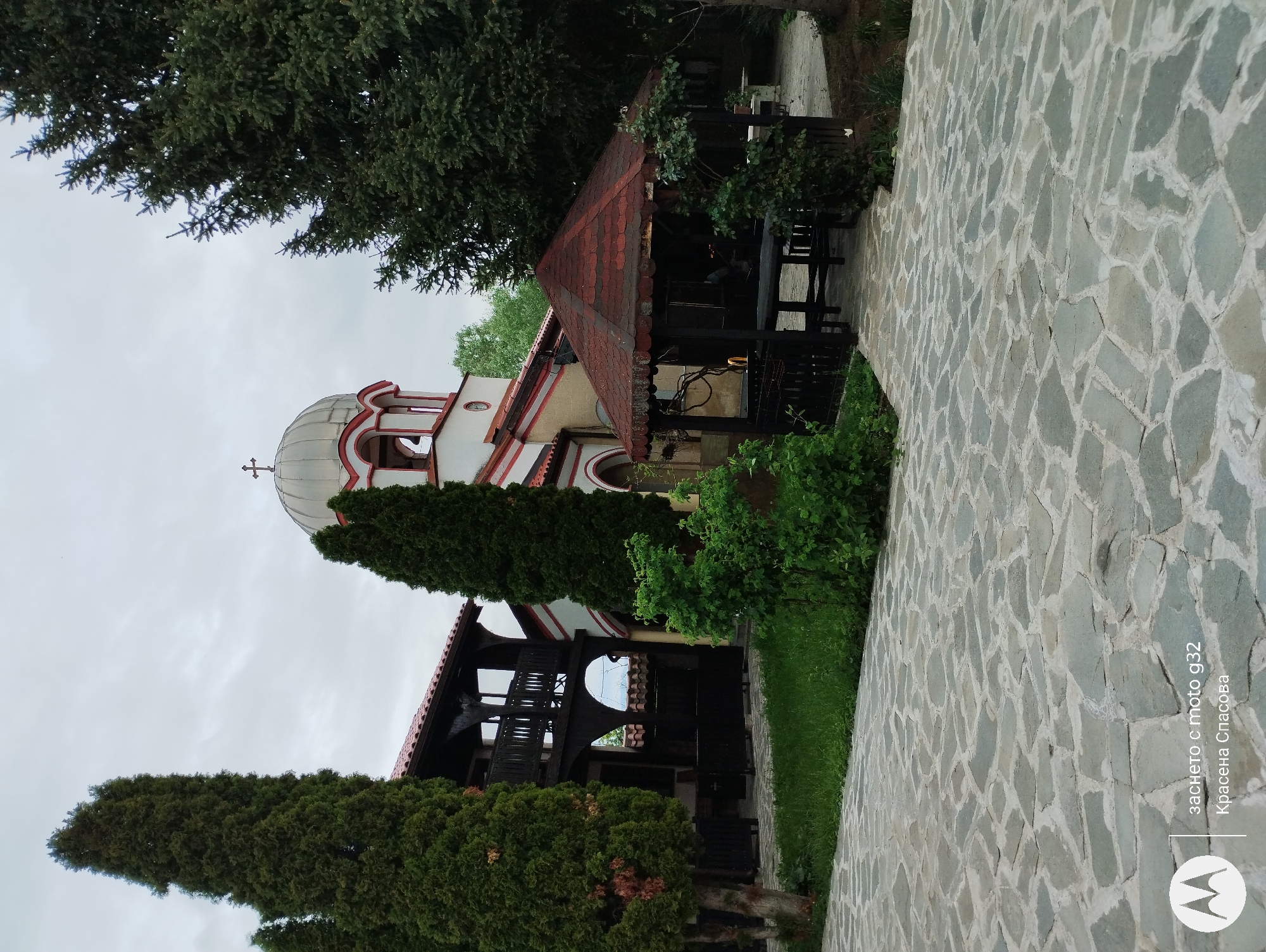 The image shows a stone path leading toward a white church with red trim in what appears to be a park-like setting. The church has a dome with a cross on top. There are tall, narrow evergreen trees flanking the church. A wooden structure with a red tile roof is located to the right of the church. The background is a cloudy sky. The path is paved with irregular gray stones. The image was taken with a Moto G32 by Krasena Spasova, but the specific location in Sofia, Bulgaria, is not discernible from the image.