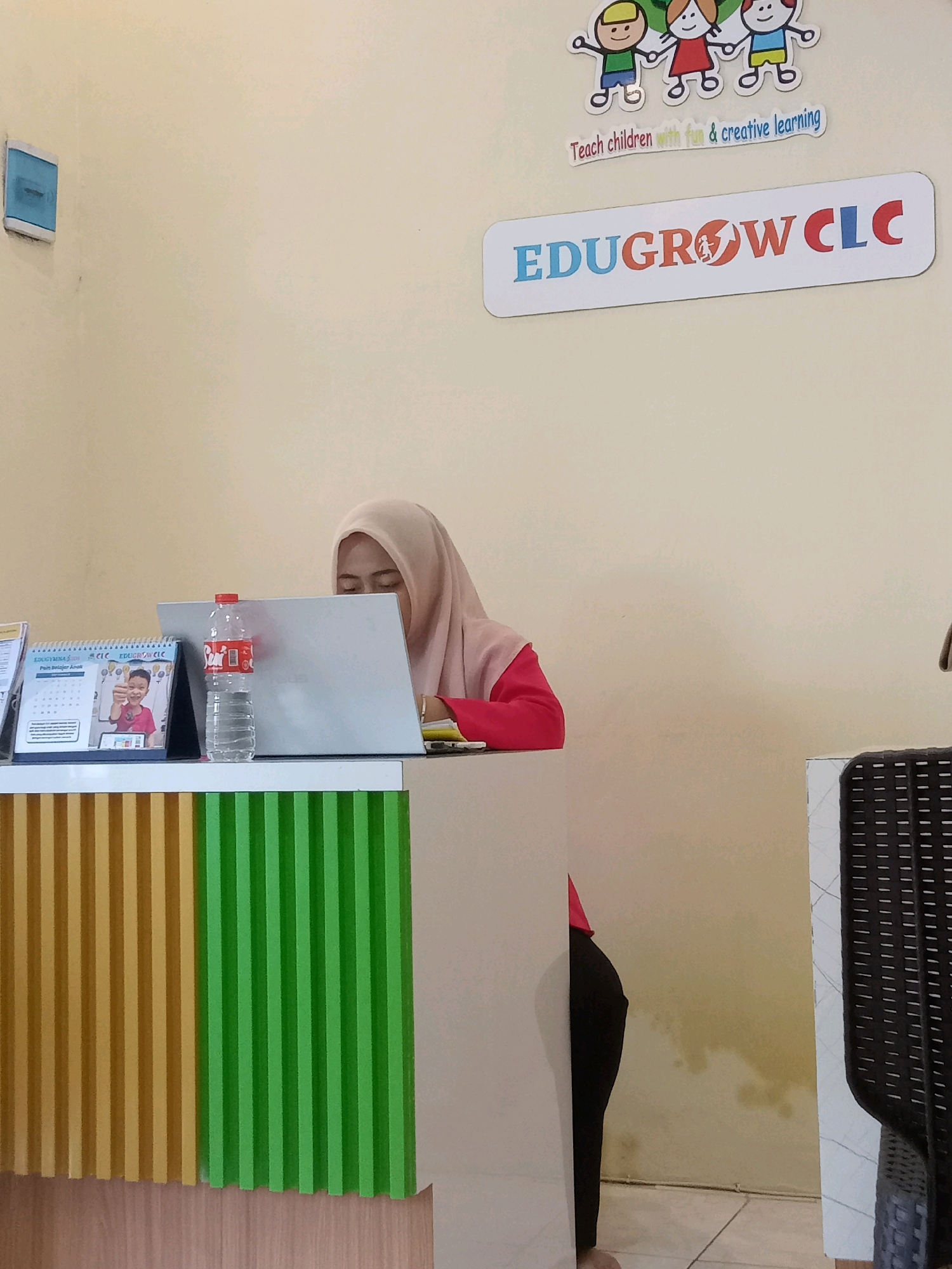 In the city of Perbutulan, Indonesia, an indoor shot captures a reception area of an educational institution. A woman wearing a light pink hijab and a pink shirt is seated behind a desk, looking down at a laptop. In front of her are a calendar and a bottle of water. The wall behind her features a colorful logo that reads "EDUGROW CLC" with a graphic of a rising arrow inside an orange circle. Above this logo, a sticker depicts three cartoon children holding hands with the text "Teach children with fun & creative learning." The reception desk itself has a vertically striped design in yellow and green. To the right of the frame, a portion of a black woven chair is visible. The overall environment suggests a clean and child-friendly educational setting.