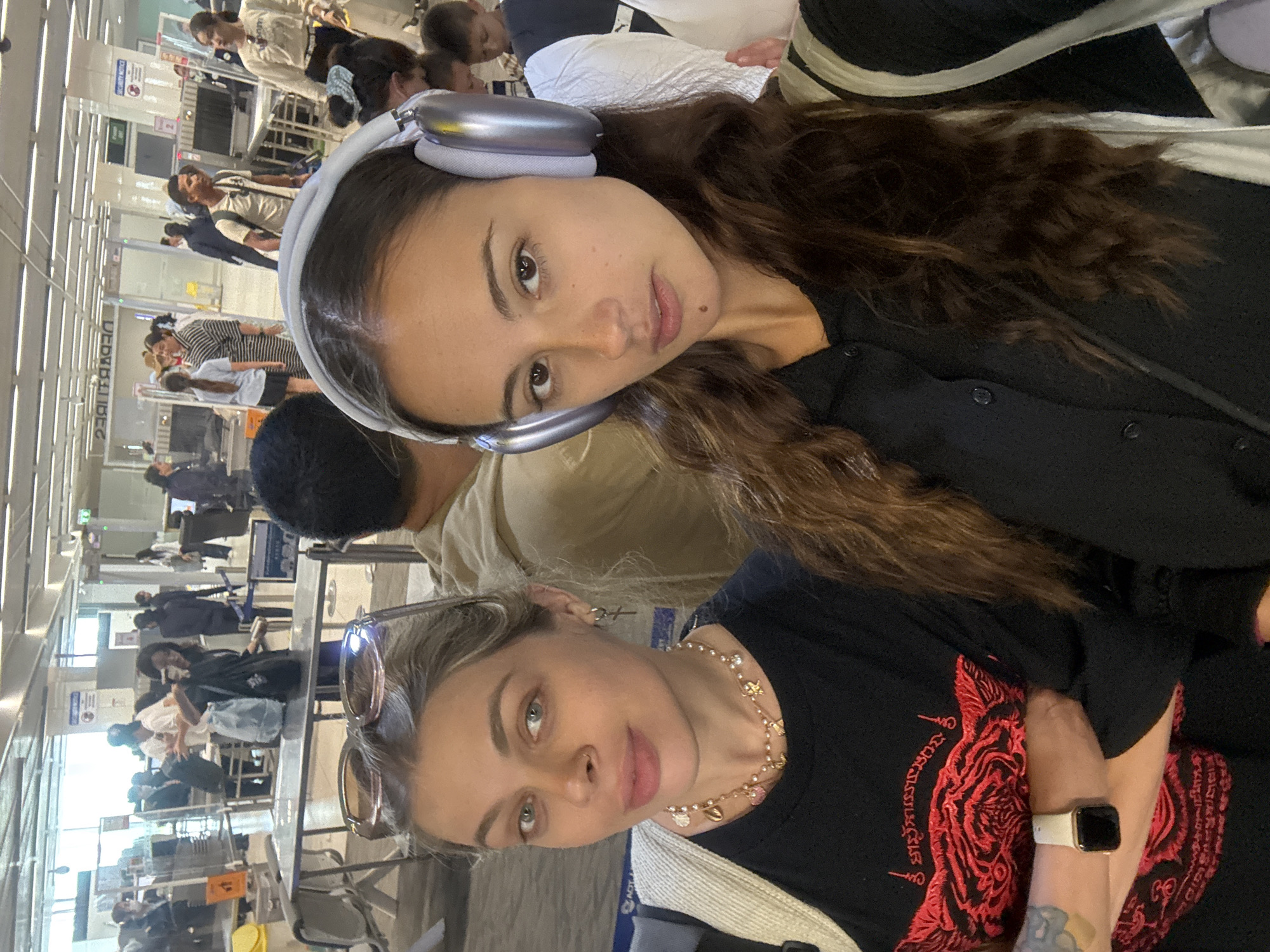 Two women are posing for a selfie at a busy airport checkpoint. The woman in the foreground is wearing a black t-shirt with red embroidery, a gold necklace, sunglasses perched on her head, and a smartwatch. The woman in the background has headphones on. Behind them is a security checkpoint with conveyor belts, bins, and numerous people lining up for screening. The image was taken in Tambon Mai Khao, Thailand.