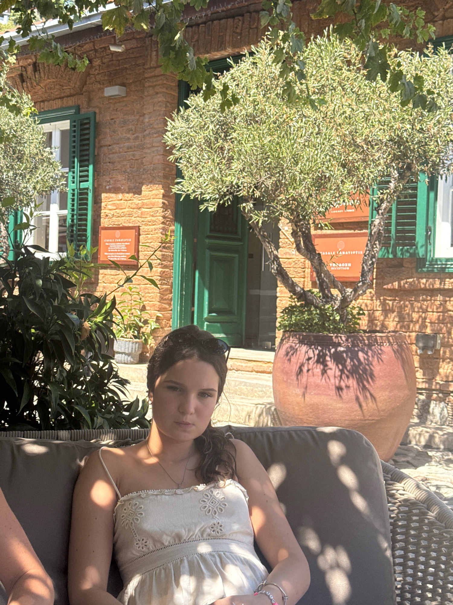 The image shows a young woman with dark curly hair, wearing a white sundress and a necklace, sitting on an outdoor sofa in what appears to be a courtyard. She is looking at the camera with a neutral expression. To her left, another person's arm is visible. Behind her, there is a traditional brick building with green shutters and doors. Several potted plants, including an olive tree in a large terracotta pot, are scattered around the area. In the background, a sign on the building reads "CASALE PANAYIOTIDES" and "RECEPTION," indicating the location as Casale Panayiotides in Kalopanagiotis, Cyprus. The scene is bathed in sunlight, casting shadows from the trees and foliage.