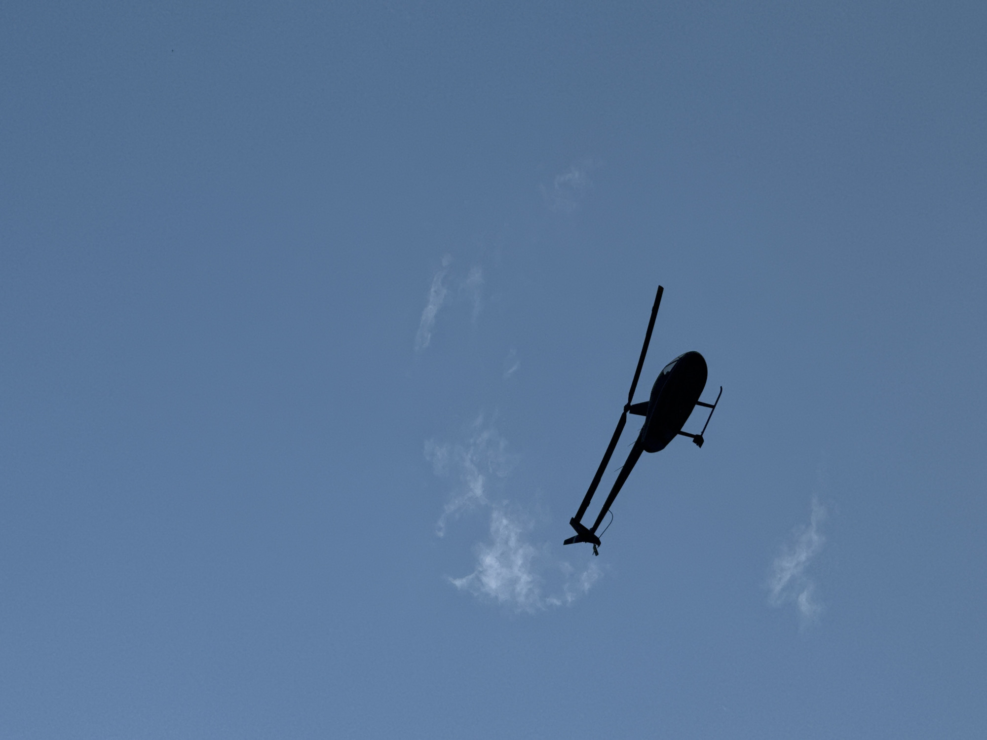 A silhouette of a small helicopter is visible in the sky, angled slightly downwards and to the right. The helicopter is dark and appears to be in flight. A few wisps of white cloud are scattered in the pale blue sky. The image captures the helicopter from below, against a clear sky. The scene is devoid of any specific landmarks that would pinpoint the location in Artybash, Russia.
