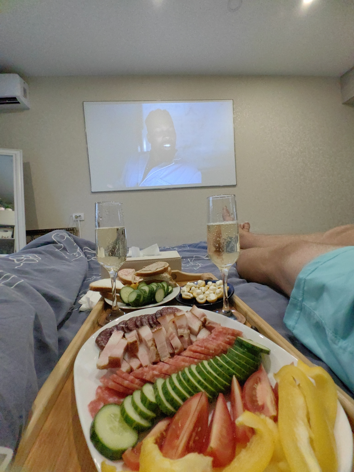 In a bedroom in Fanipal, Belarus, a person lies in bed, covered by a blue duvet with a white line drawing of clouds. They are watching a projected movie on the wall, visible on a large white screen. On a wooden tray placed on the bed is a breakfast spread. Two glasses of champagne stand next to a plate of sliced meats, including salami and bacon, and a variety of vegetables such as sliced cucumbers, tomatoes, and yellow bell peppers. There are also small sandwiches and a dish of what appears to be cheese bites. The person's legs are visible in the frame, indicating they are likely a man wearing blue shorts. The room appears to be illuminated by the light from the projected movie. The overall mood is relaxed and intimate, suggesting a leisurely morning or evening spent enjoying a movie and a meal in bed.