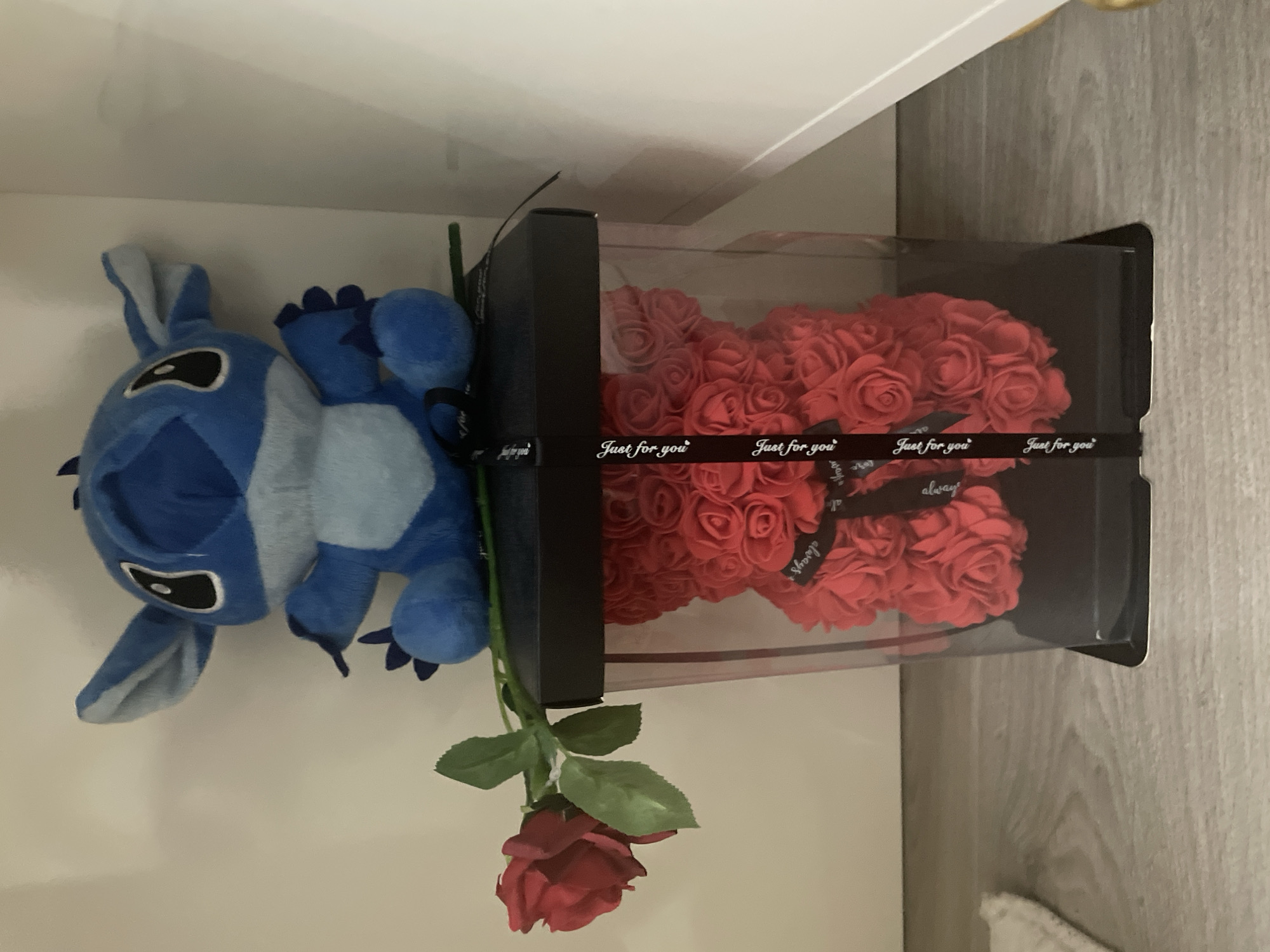 In Ihtiman, Bulgaria, an indoor scene displays a blue plush Stitch toy positioned on a black rectangular box. A single artificial red rose with green leaves lies horizontally across the top of this black box. Below, a transparent square display case, featuring a black base and lid, contains a red rose bear composed entirely of numerous small red artificial roses. A black ribbon, printed with repeating white text "Just for you," is wrapped vertically around the transparent case. The objects are situated on a light-colored wood-grain surface. A plain light-colored wall forms the background. To the lower left, a partial white fluffy textile object is visible, and to the lower right, a white vertical panel with a gold-colored accent is present.