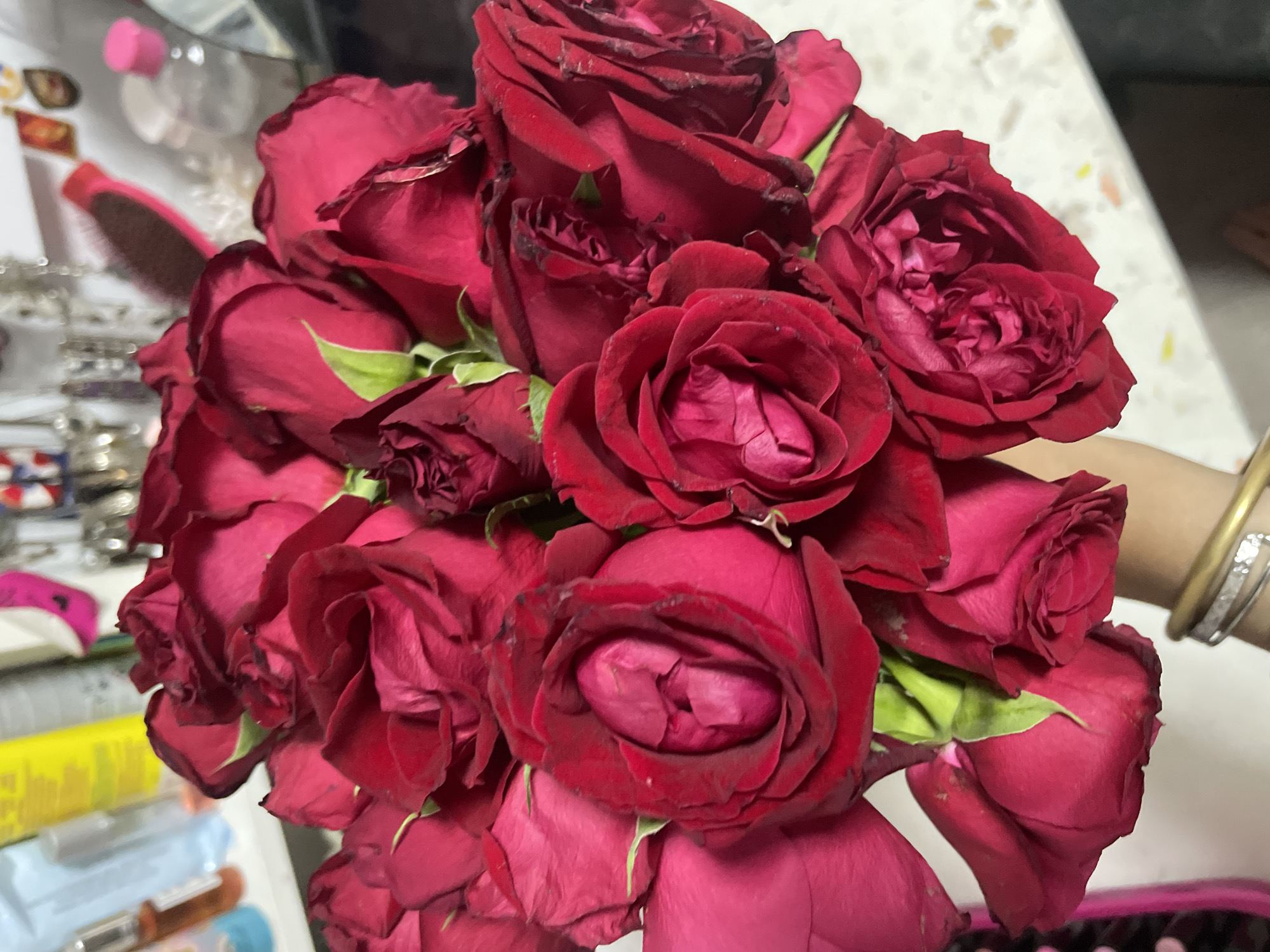 A close-up, overhead shot shows a bouquet of deep red roses. The roses are in various stages of bloom, with some fully open and others still tightly furled. The petals are velvety and rich in color, with subtle variations in shade and texture. Some of the petals have slightly dry or browned edges, indicating they may not be freshly cut. Green stems and leaves are visible in places, providing a contrast to the dark red of the blooms. The background is blurred but suggests an indoor setting with some brightly colored objects, possibly toiletries or cosmetics, and what appears to be a white, speckled surface. The image is taken from a high angle, looking down onto the arrangement of flowers. There is no indication of the location being in Raanana, Israel.