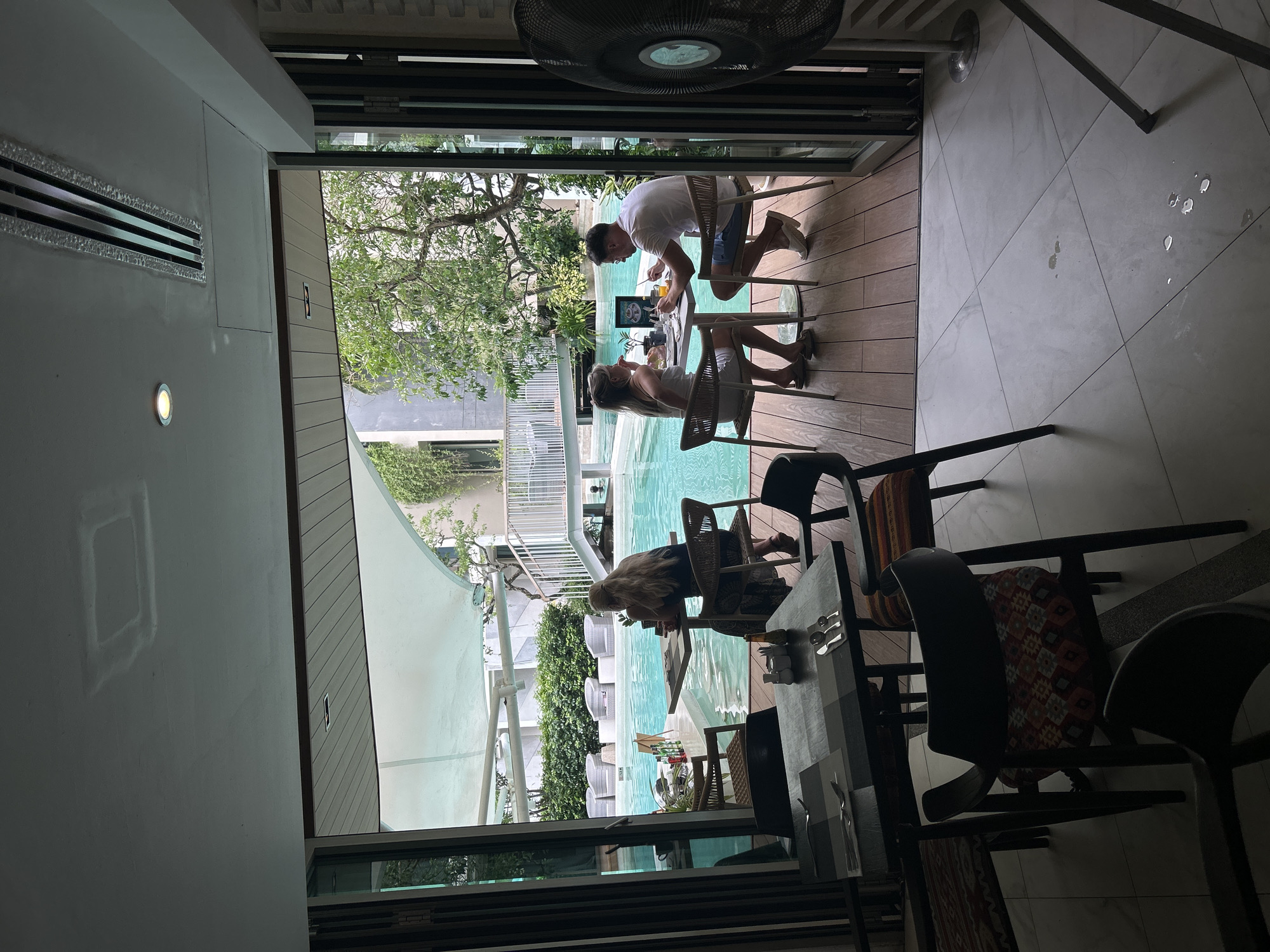 The photograph depicts an indoor-outdoor restaurant or cafe setting, likely in Rawai, Thailand, judging by the presence of a swimming pool in the background. Three individuals are seated at tables near an open-air portion of the establishment. Two of them are conversing over a meal at one table, while the third person is at a separate table and is looking at their phone. The seating area overlooks a swimming pool with manicured greenery and building structures in the background. The foreground of the image includes interior elements such as tables and chairs, with a floor fan partially visible in the upper section of the frame. The overall lighting suggests daylight hours.