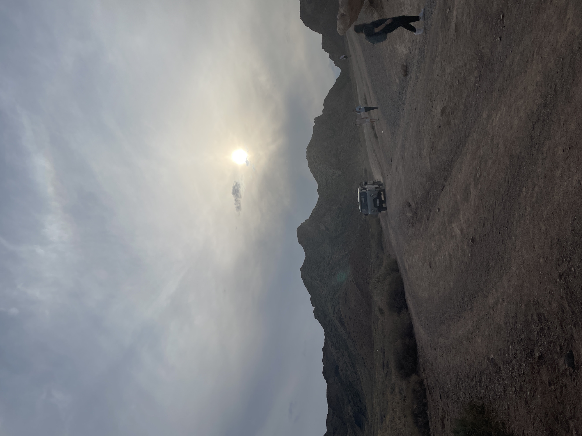 The image depicts a rugged, arid landscape with rolling hills and mountains under a hazy, overcast sky where the sun is partially obscured, creating a diffused glow. A dirt road winds through the scene, with tire tracks visible. Several people are present: one individual in the foreground on the right, facing away from the camera and wearing a black top, dark pants, and white sneakers with a backpack; two individuals walking up the road towards the center of the frame, one wearing a light-colored top and shorts, the other a blue top and darker pants; and another person further up the road. A light-colored SUV is parked on the left side of the road, facing uphill. The vegetation is sparse, consisting of dry brush and low-lying plants. The overall environment suggests a natural, undeveloped area, consistent with the outskirts of Tamerlik, Kazakhstan, known for its natural parks and mountainous terrain.