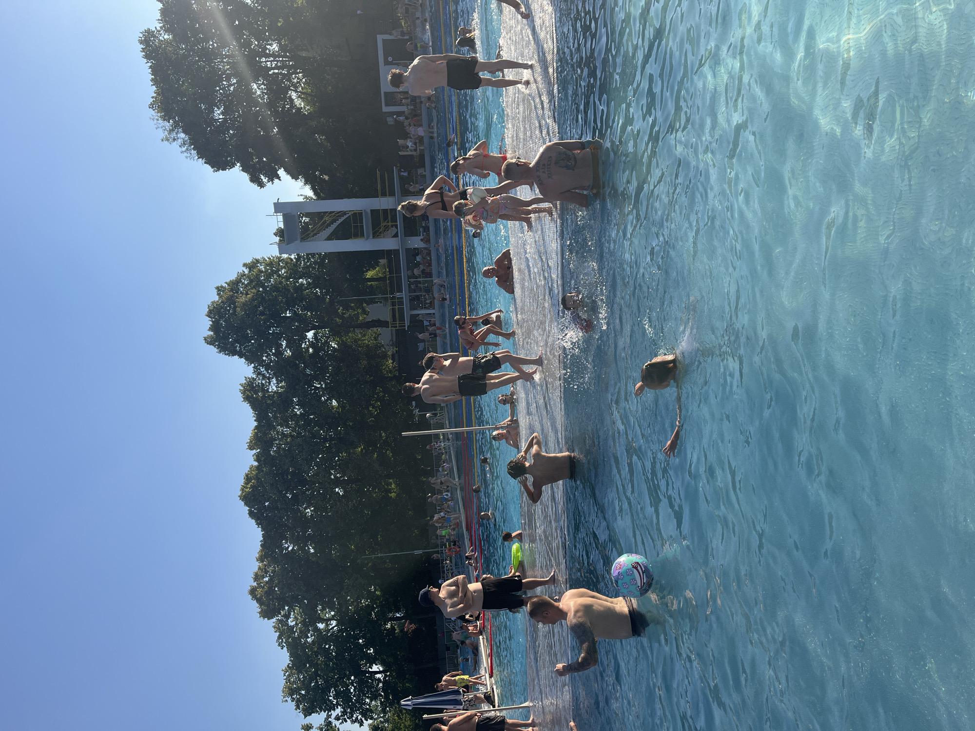 This media file captures a bustling outdoor swimming pool on a sunny day. The scene is filled with people enjoying various aquatic activities. Several individuals are in the water, some swimming, others standing or wading. A few people are positioned on the edge of the pool, either preparing to enter or resting. One person is in the process of diving or jumping into the pool from a raised platform visible in the background.
Objects present include a colorful inflatable ball floating in the water, suggesting playful interaction. The pool itself is a bright blue, with clear water indicating good maintenance. The surrounding area features lush green trees, providing shade and a natural backdrop. In the distance, parts of a diving tower and other swimming lanes are visible, implying a larger aquatic facility.
The time of day appears to be midday or early afternoon, judging by the bright sunlight casting shadows and illuminating the water. The clear blue sky suggests fair weather. The overall mood of the scene is lively and recreational, with people engaged in leisure activities. There is no visible text in the image.