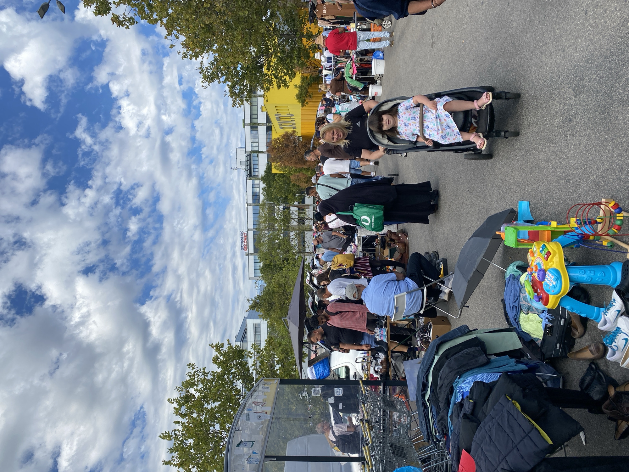 The image depicts an outdoor market scene in Vienna, Austria. The market appears to be a flea market or a similar vendor event, with numerous stalls and people browsing. Various goods are displayed, including clothing, shoes, and toys. In the foreground, a child is seated in a stroller, pushed by an adult. Several other adults are visible, some interacting with vendors or examining merchandise, while others are walking through the market. The background shows buildings and trees, indicating an urban setting. The sky is partly cloudy.