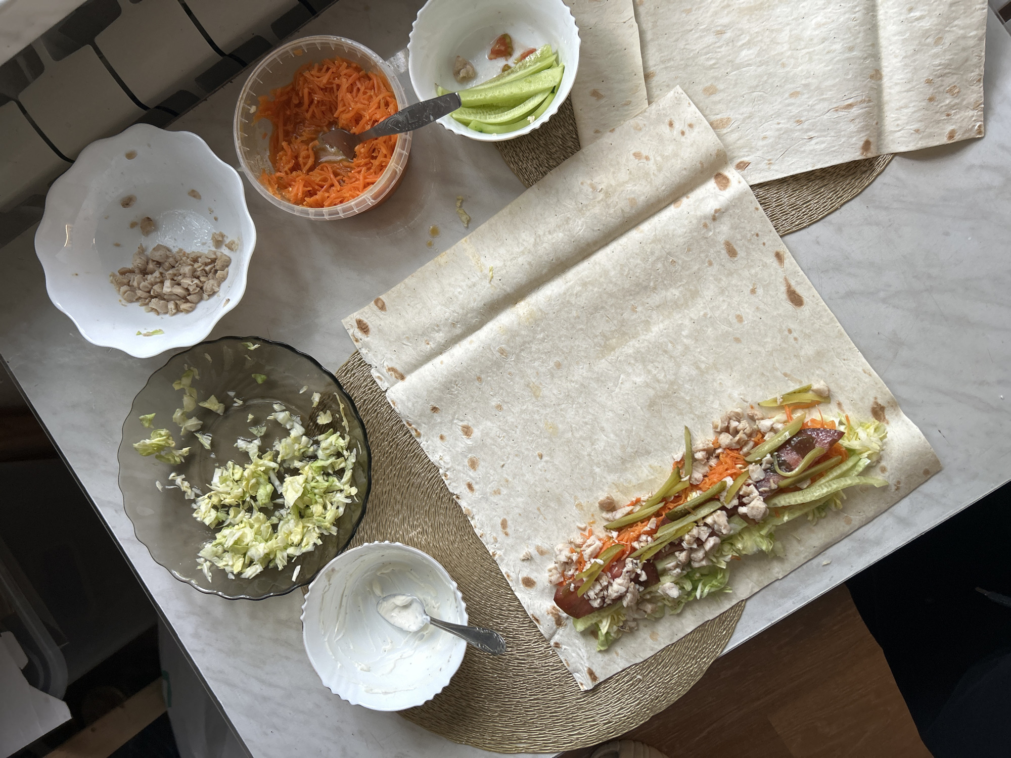 This overhead shot captures a scene of food preparation on a light-colored countertop, likely in a kitchen. The primary subject is a large piece of lavash bread, partially rolled and filled with various ingredients. These ingredients include shredded lettuce, grated carrots, chopped chicken, slices of cucumber, and what appears to be a piece of sausage or a similar cured meat. A small bowl of white sauce, possibly mayonnaise or sour cream, with a spoon resting in it, is visible to the left of the lavash. Nearby, a clear plastic container holds more grated carrots, and a white bowl contains sliced cucumbers. Another dark glass plate is filled with chopped lettuce.
The overall impression is one of a home-cooked meal being assembled. The lighting suggests it might be daytime, with natural light potentially coming from a window not visible in the frame. There are no people visible in the image. The setting is functional, with a focus on the food preparation process. The items are arranged on a woven placemat, adding a touch of rustic texture to the scene. The chopped chicken suggests the meal being prepared is a type of wrap or shawarma.