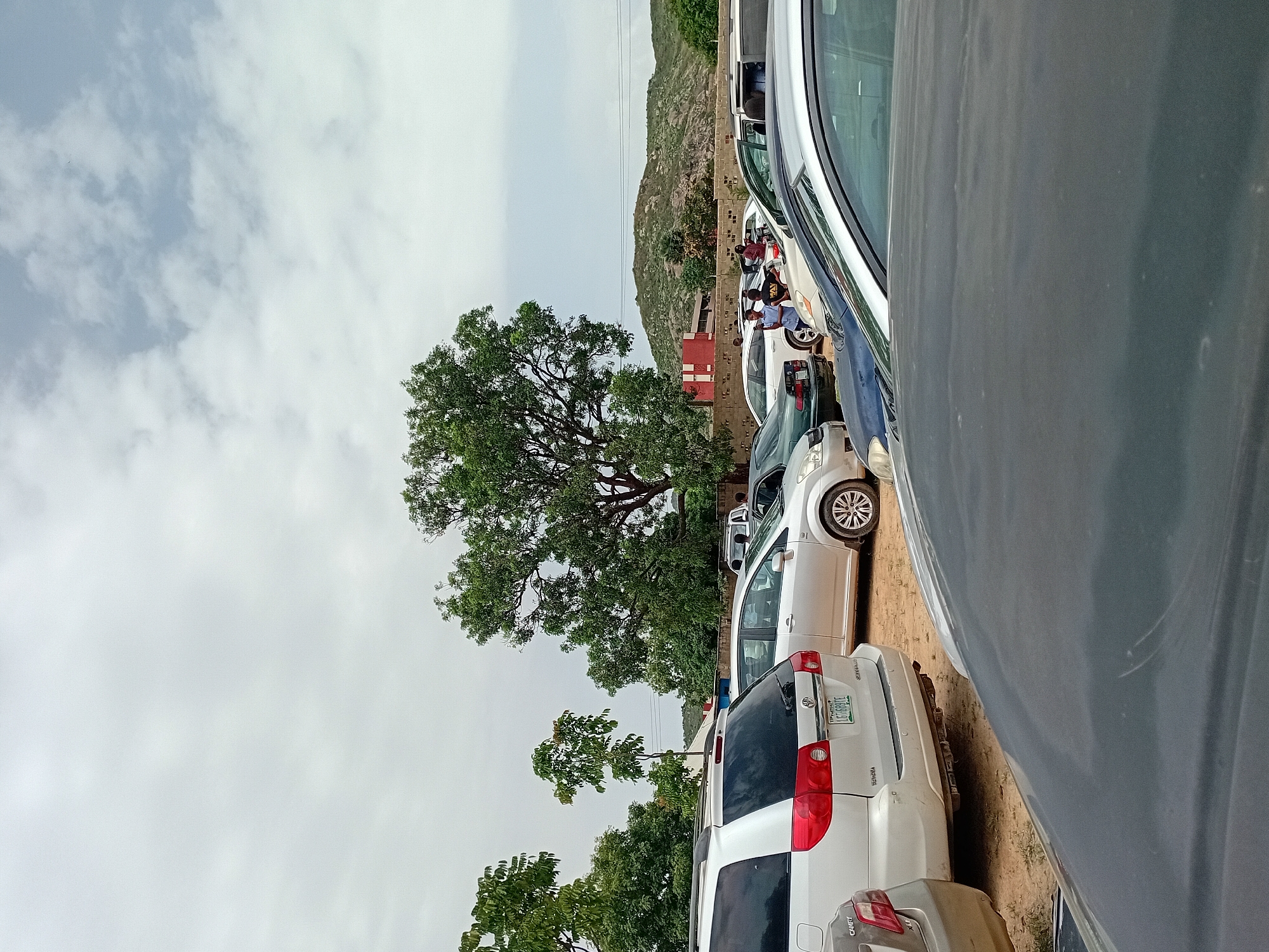 The image shows a parking lot scene in Jos, Nigeria. Several vehicles are parked closely together, primarily white and gray cars and minivans. In the background is a rocky hill, and a red-roofed building is partially visible. Several people are gathered near one of the vehicles. A large tree is situated near the middle of the scene. The sky is overcast with heavy cloud cover. Powerlines are also visible in the sky. The parking lot appears to be on a dirt or gravel surface.