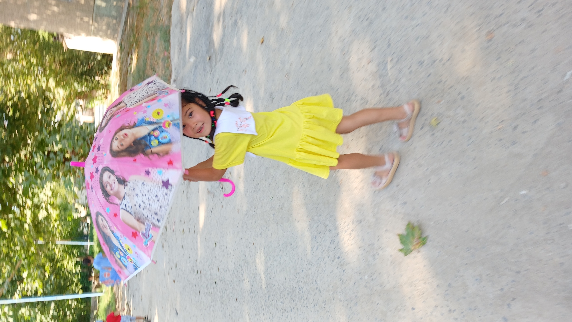 A young girl in a yellow dress and sandals is holding an open pink umbrella decorated with pictures of girls. She is walking on a paved path outdoors, with trees and greenery in the background. The scene appears to be in a park or recreational area. The image is blurry, suggesting motion.