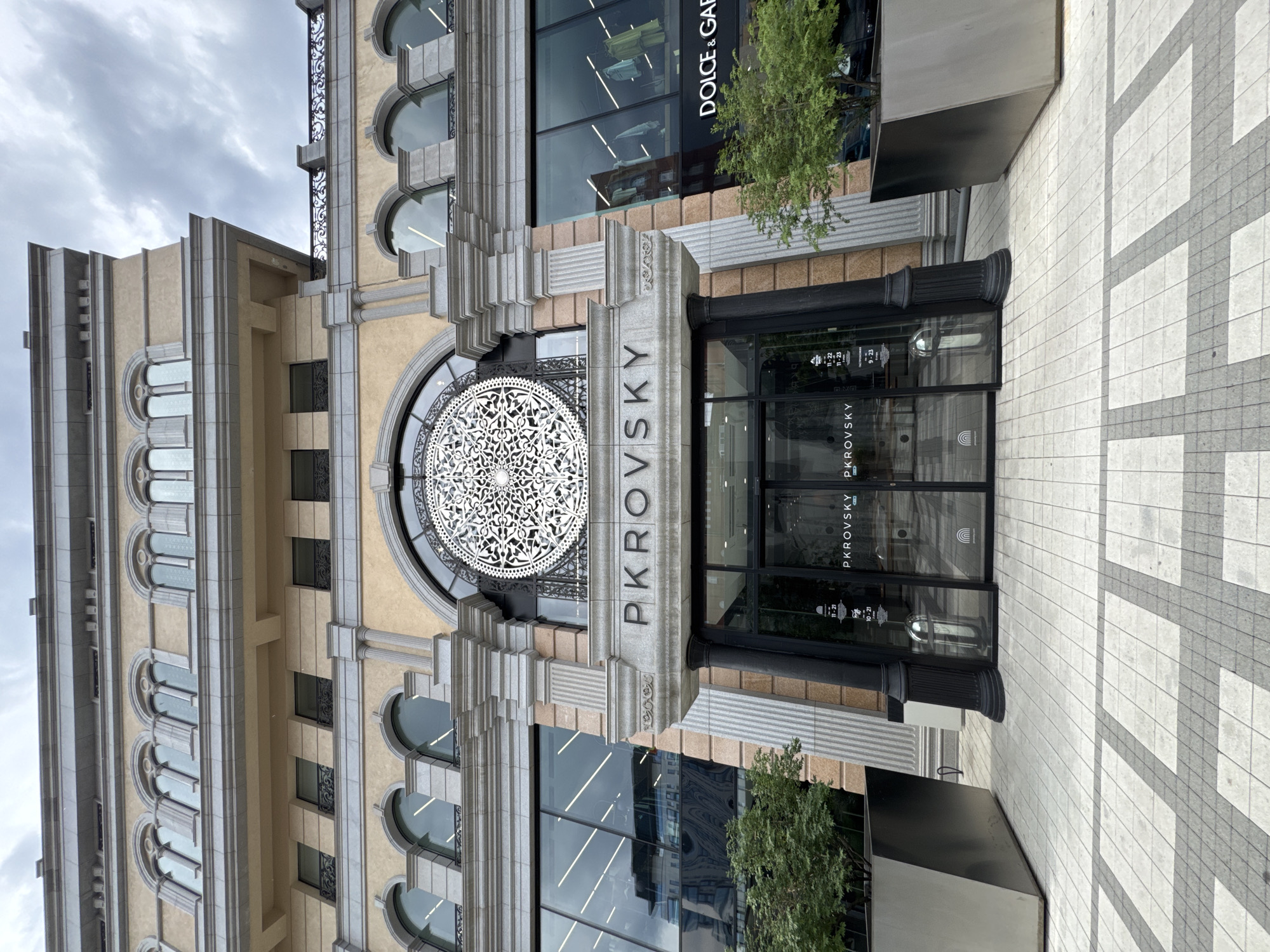 This is a vertical, eye-level, medium shot of the facade of the Pkovsky store in Yekaterinburg, Russia. The building is ornate with a cream-colored stone facade, intricate carvings, arched windows, and decorative metalwork. A large, circular, white decorative metal medallion is centered above the entrance. The entrance itself is a modern glass-fronted doorway with "Pkovsky" written vertically on the glass. Above the entrance, the word "Pkovsky" is also carved into the stone in large capital letters. To the right of the entrance, a sign is visible on the glass, displaying store hours. A small, green tree is planted to the right of the entrance. The sidewalk in front of the store is paved with light and dark gray square tiles arranged in a geometric pattern. The sky above is overcast and appears to be a cloudy day. The overall impression is of a grand and historic building with a modern retail establishment.