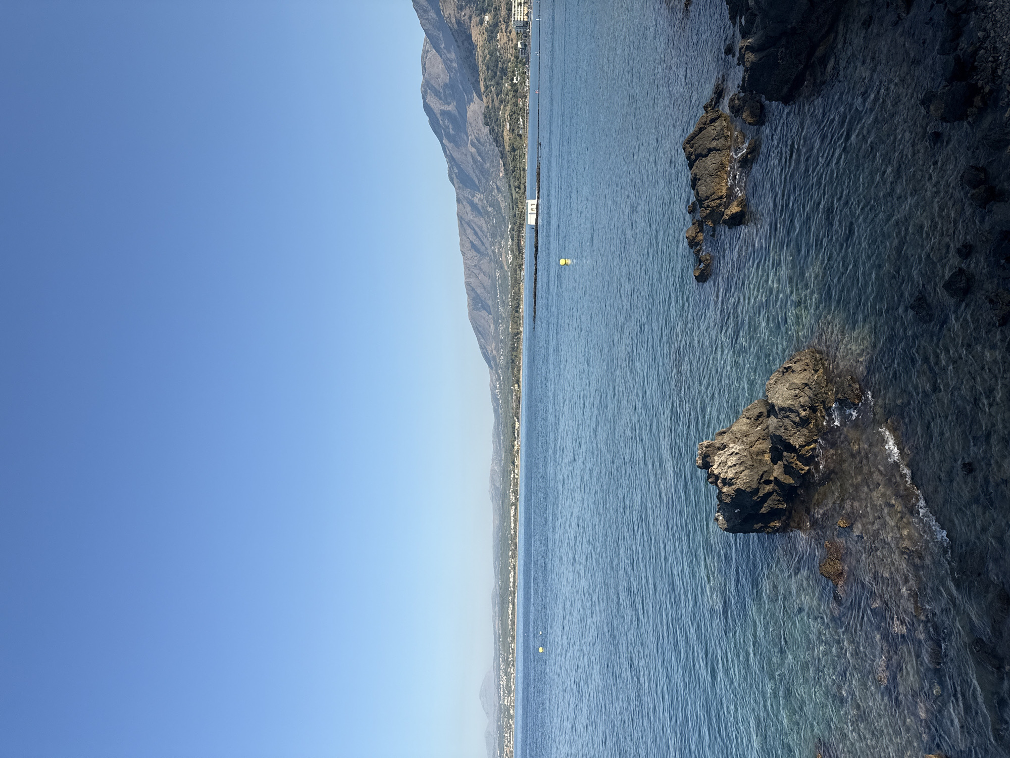 The image depicts a serene coastal scene in Georgioupoli, Greece. The foreground features rocky outcrops emerging from the clear, blue water, revealing the seabed beneath. In the mid-ground, the calm sea stretches towards a distant shoreline, dotted with buildings and a prominent white chapel situated on a small island connected by a causeway. Mountains rise in the background, forming a natural amphitheater around the bay. The sky is a clear, uninterrupted blue, indicating a sunny day. There are no people clearly visible in the scene, though distant figures may be present along the shore. The overall atmosphere is tranquil and picturesque, characteristic of a Mediterranean seaside town.