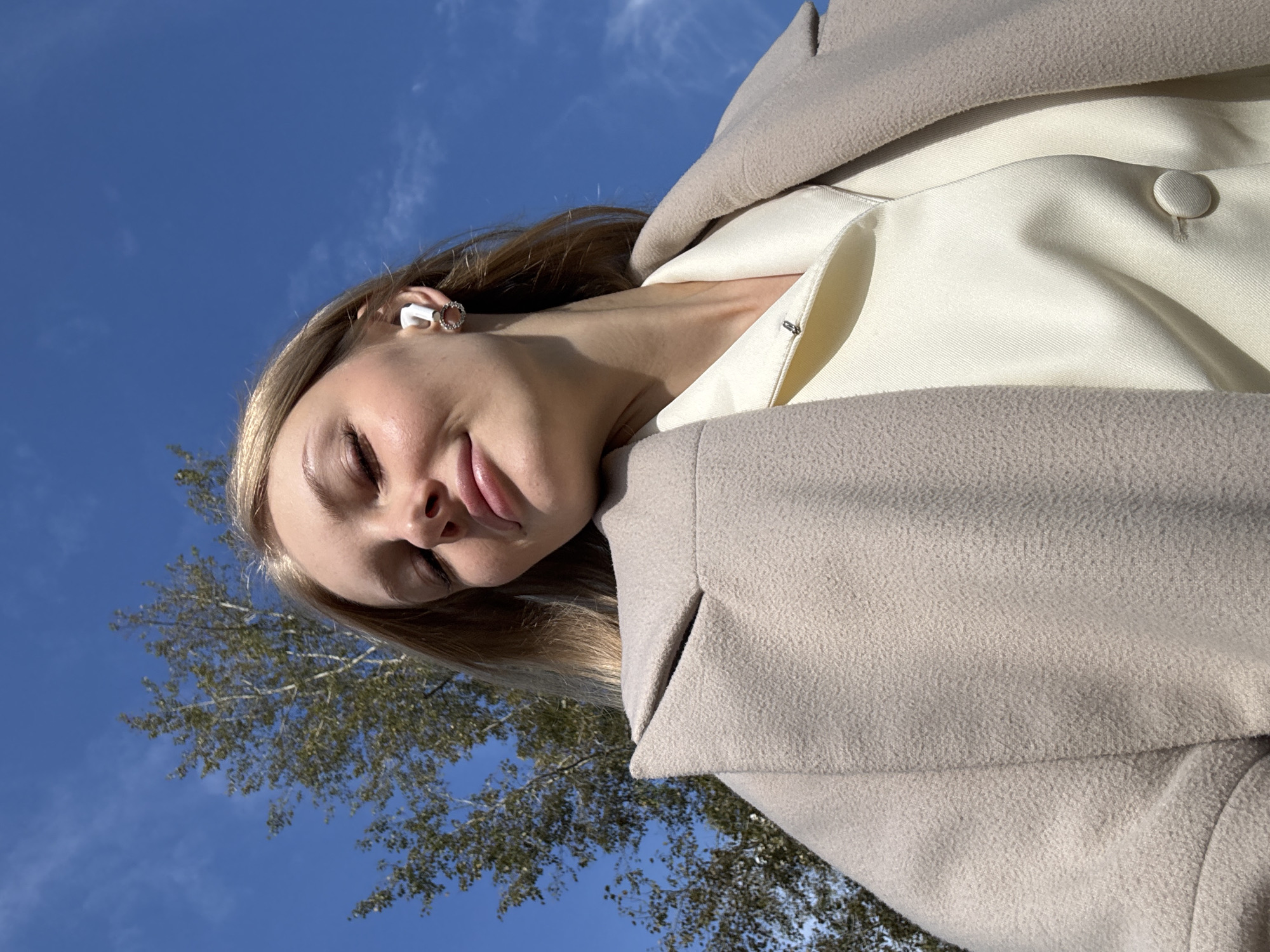 In the city of Perm, Russia, a young woman with blonde hair and closed eyes poses for a selfie outdoors against a bright blue sky and a distant tree. She is wearing a cream-colored coat over a matching blouse, and a white wireless earbud is visible in her right ear. Her expression is serene, with slightly parted lips. The lighting suggests it is daytime.