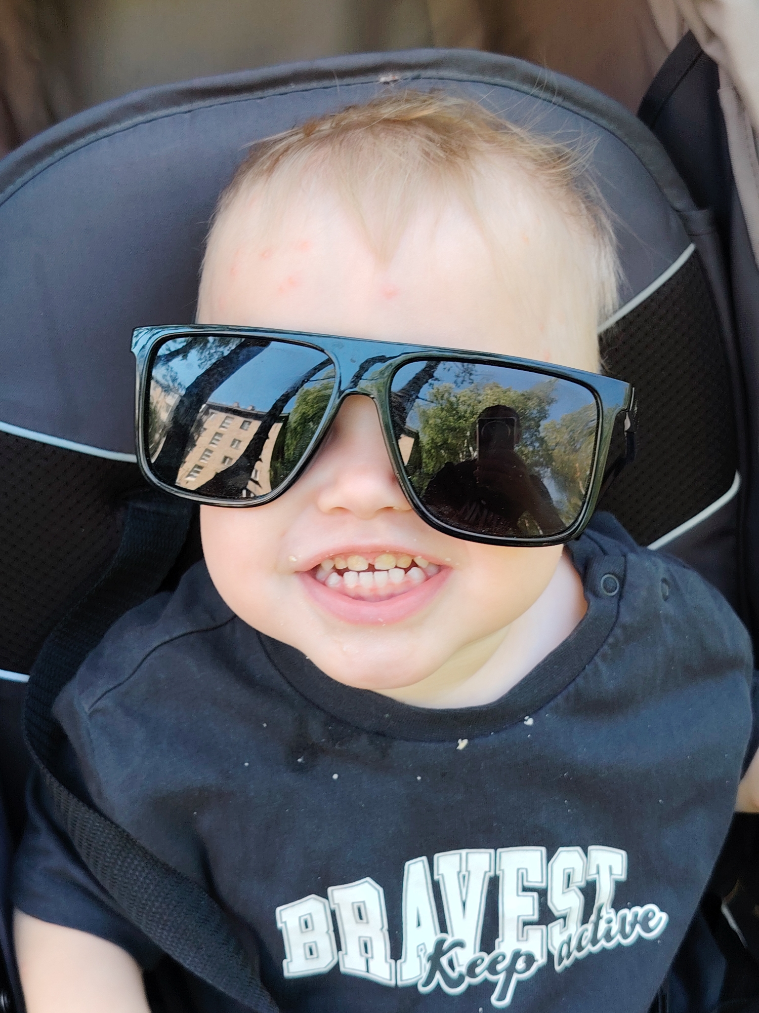 A young child wearing oversized black sunglasses and a black t-shirt with the word "BRAVEST" printed on it is sitting in a stroller. The child has fair hair and a rash on the forehead. The sunglasses reflect a building and trees. The location is suggested to be Chaikovsky, Russia, based on the building's architecture reflected in the sunglasses. The child appears to be smiling, exposing a few teeth. Crumbs are visible on the child's t-shirt and around the mouth. The stroller is black and grey. A portion of a seat belt strap is visible across the child's chest. A person is reflected in the sunglass.