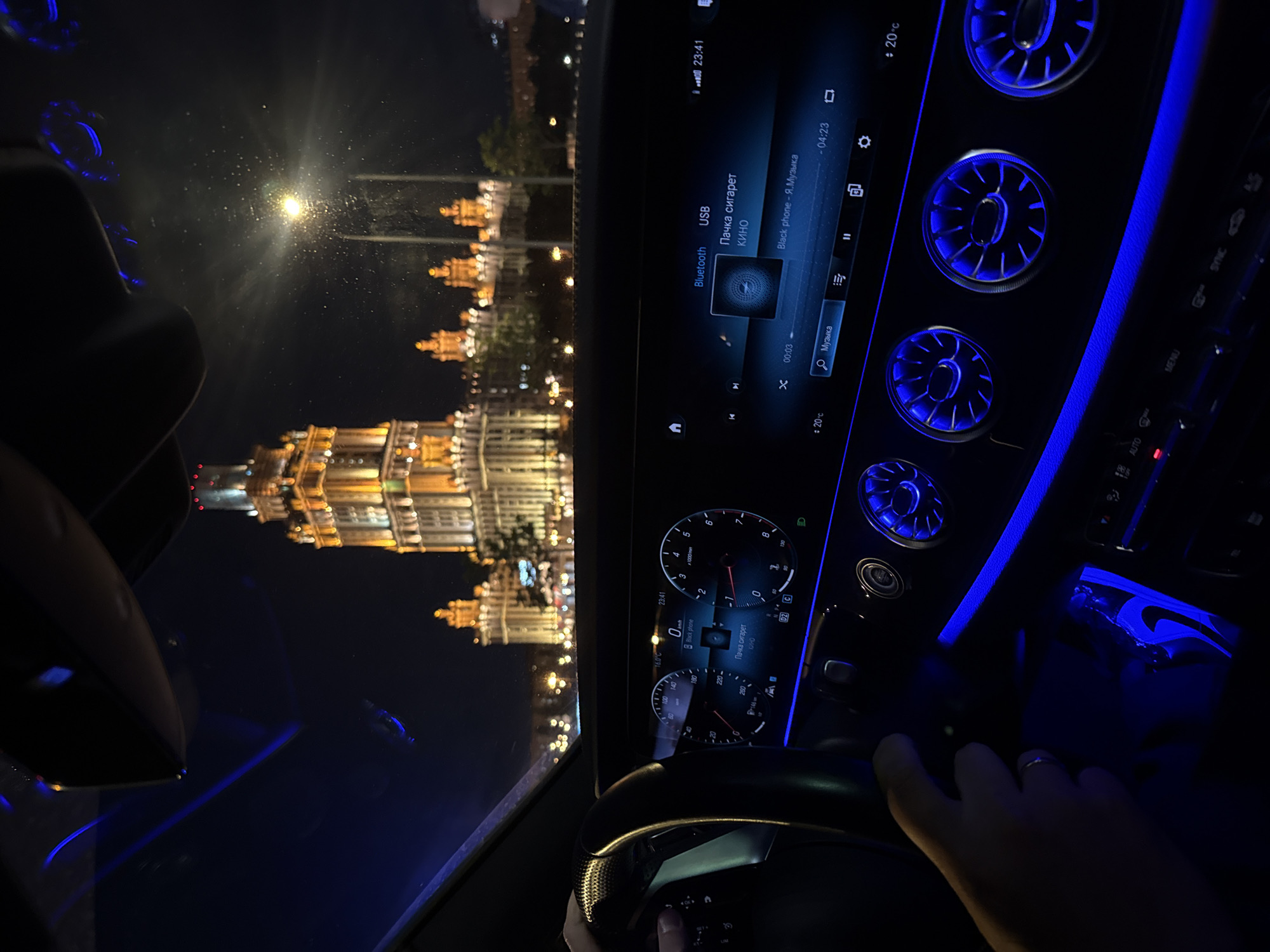 The image shows a nighttime view from inside a car, looking towards a brightly lit building in Moscow, Russia. The building appears to be an ornate, multi-story structure with a distinctive tower, reminiscent of Stalinist architecture, likely one of the Seven Sisters skyscrapers. The car's interior is illuminated by blue ambient lighting, highlighting the steering wheel, dashboard, and air vents. The dashboard displays a digital speedometer, tachometer, and an infotainment screen showing music playback information, including the song title "Пачка сигарет" by "КИНО" and "Black phone" by "Я.Музыка." A person's hand, wearing a ring, is visible on the steering wheel, indicating they are driving. The windshield is speckled with water droplets or debris, and the streetlights outside reflect on its surface. The overall scene depicts a driving experience at night in a major city, with the iconic Moscow architecture serving as a backdrop.