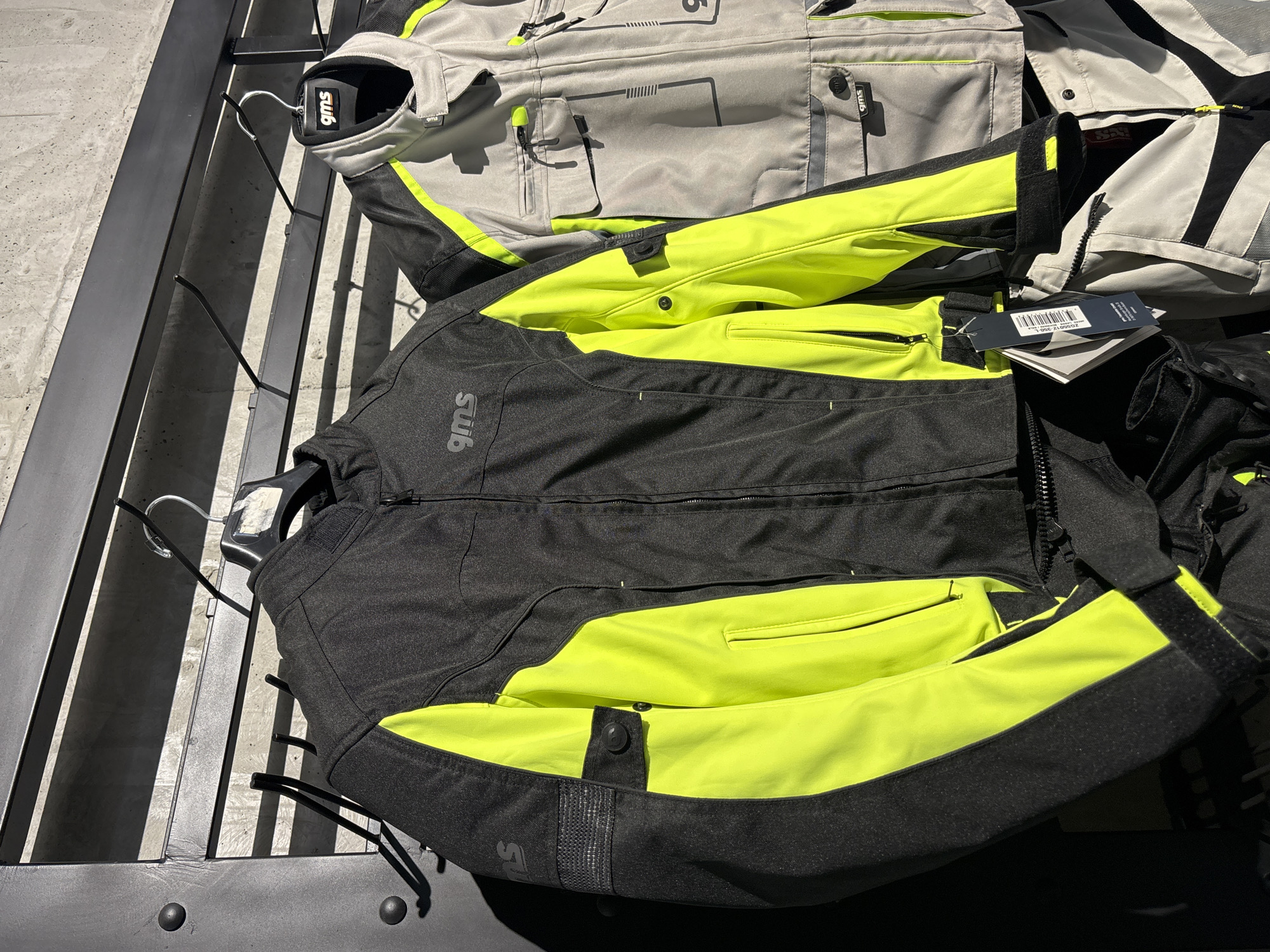The image shows a close-up of two GMS brand motorcycle jackets hanging on a rack against a concrete wall. The jacket in the foreground is black with bright neon yellow accents on the sides of the torso and sleeves. It has a zippered front closure, multiple pockets, and reflective detailing on the upper arm. The second jacket, partially visible behind the first, is light grey with similar neon yellow accents and multiple chest pockets. Both jackets are on hangers and appear to be new, with tags attached to the front jacket. The lighting suggests an indoor setting with natural light casting shadows from the rack onto the wall. The scene is devoid of people. There is no identifiable information within the image to suggest a location in Tbilisi, Georgia.