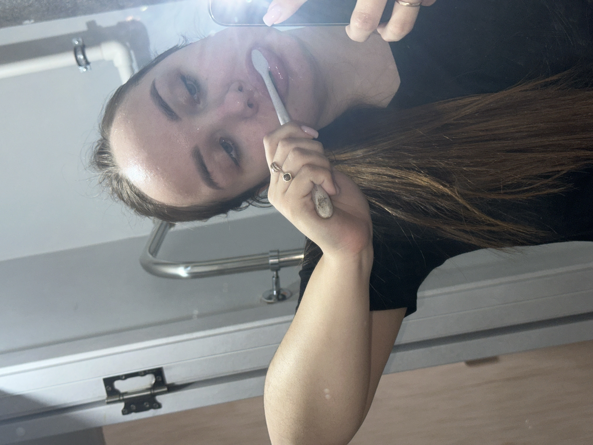 A young woman is holding a toothbrush in front of her mouth, as if she is brushing her teeth, while she is looking at her phone. The location appears to be in a bathroom. It is not possible to determine the specific location in Chaikovsky, Russia, from this image.