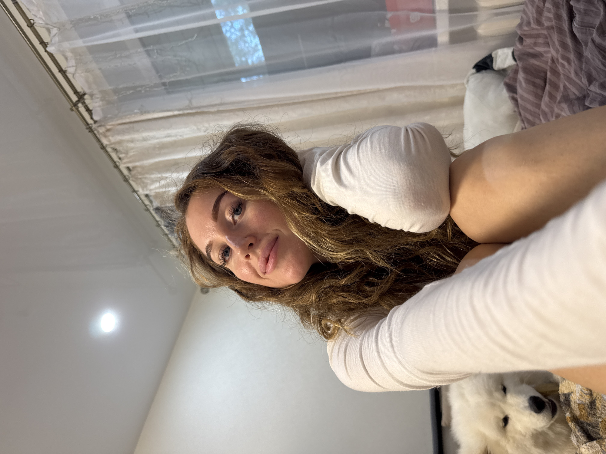 An indoor selfie captures a young woman and a fluffy white dog, likely a Samoyed. The woman, positioned in the lower-left, has long, wavy brown hair, blue eyes, and a gentle smile, looking directly at the camera. She wears a plain white long-sleeved top, and her arm is extended, holding the camera. The white dog's head is partially visible in the lower-right, resting near the woman's arm.
The background of the image reveals a domestic setting. A window with sheer white curtains is visible on the left side, indicating natural light and suggesting it is daytime. On the right, a light-colored ceiling features a distinct, bright, circular artificial light source. Further right, a patterned fabric in tones of brown and yellow, and a striped purple and grey fabric are visible. The overall mood is relaxed and comfortable. There is no visible text in the image. The setting is an interior, located in Chaikovsky, Russia.
