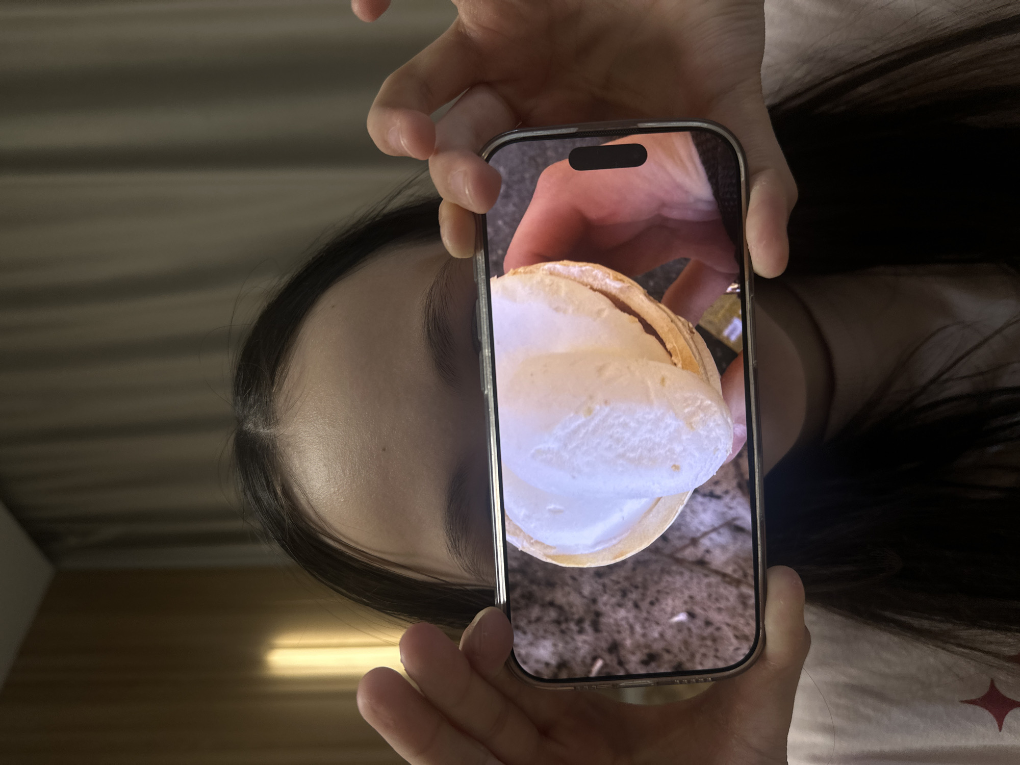 An overhead, close-up shot captures a person holding a silver-colored smartphone, likely an iPhone given the visible Dynamic Island notch. The person's hands grip the phone, which displays a picture of a dessert: a scoop of white, creamy substance (possibly ice cream or whipped cream) served in a natural, light-brown shell, resembling a coconut. The background of the phone screen image shows a textured, dark surface.
Parts of the person's face, including an eyebrow, eye, and forehead, are visible on the left side of the frame, indicating dark hair. The background behind the person is out of focus, featuring horizontal lines and a soft, warm light source on the lower right, suggesting an indoor setting. The image's orientation implies the person may be reclining or lying down. No discernible text is visible on the phone or in the surrounding environment. The context for this media file is Votkinsk, Russia.
