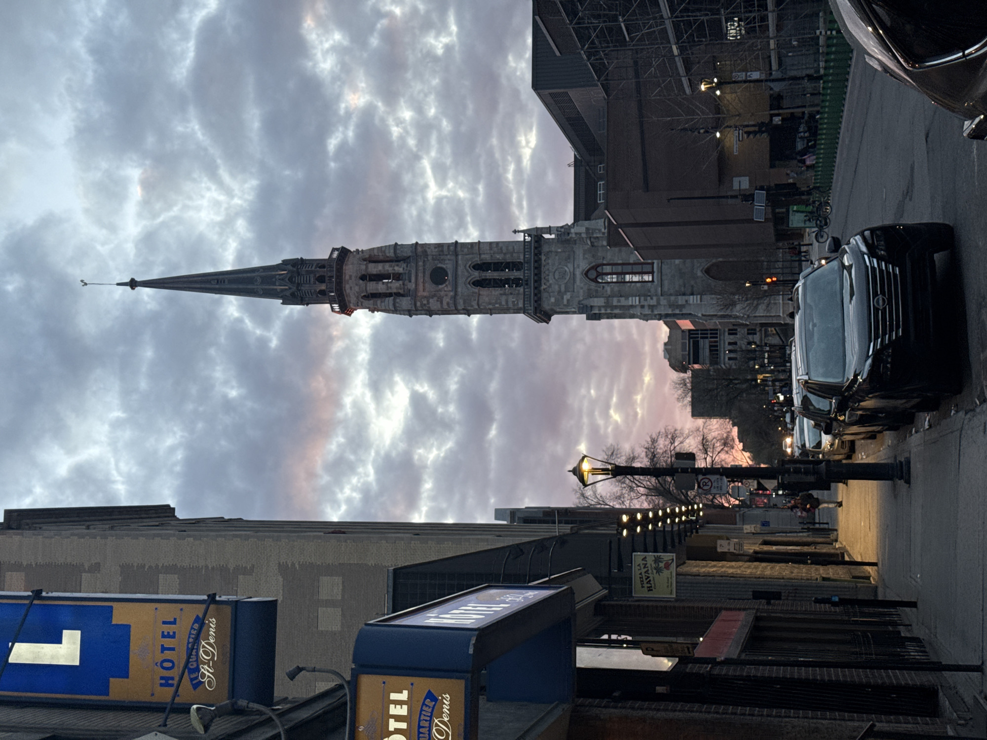 This photo depicts a Montreal street scene at dusk. Dominating the frame is the steeple of the Saint-Patrick's Basilica. The street is lined with buildings, streetlights, and parked cars. Hotel signs with the wording "Hotel Quartier St-Denis" are seen on the left. The sky is cloudy, with hints of pink. The street appears relatively empty, with only a few distant figures visible.