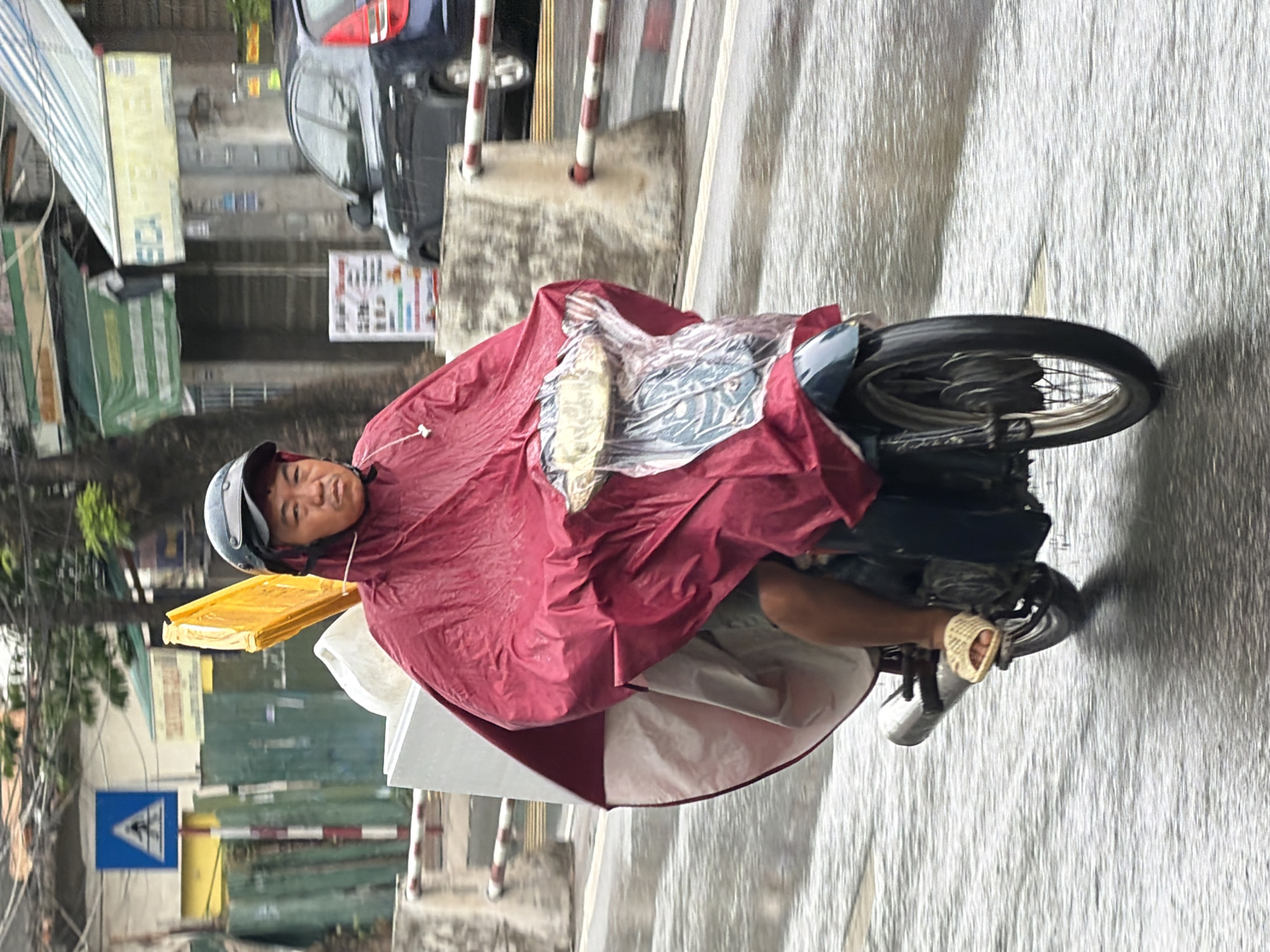 A person on a motorcycle wears a large red poncho and a black helmet. The poncho covers the person and a large, light-colored box strapped to the back of the motorcycle, as well as a yellow, rectangular object protruding from the top of the box. The person is wearing light-colored woven sandals. The motorcycle has a chrome exhaust pipe and is on a wet, paved road, indicating it is raining. In the background, there are buildings with awnings, a black car, and traffic barriers. A blue and white road sign is visible on the left side of the image. The scene appears to be on a street in Nha Trang, Vietnam, given the context of the location.