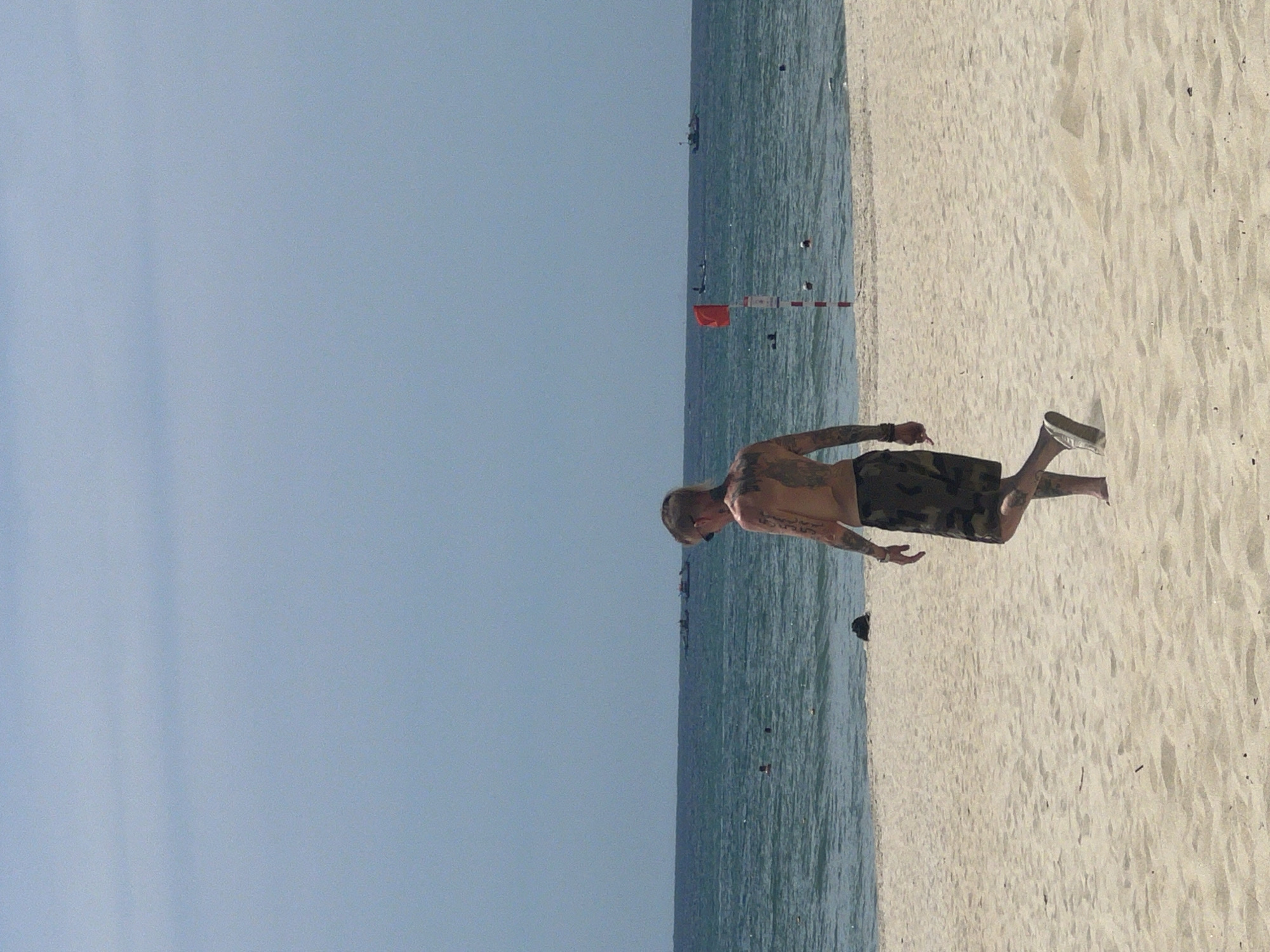A shirtless man with extensive tattoos on his back and arms, wearing camouflage-patterned shorts and white sneakers, walks away from the camera on a sandy beach. He is in the foreground of the image. The sea is visible in the background, with several small boats and a few people swimming in the water. A red flag on a pole stands in the middle ground of the sea, indicating a hazard. The sky is clear and blue. The overall setting appears to be a beach in Đà Nẵng, Vietnam, likely during daylight hours.