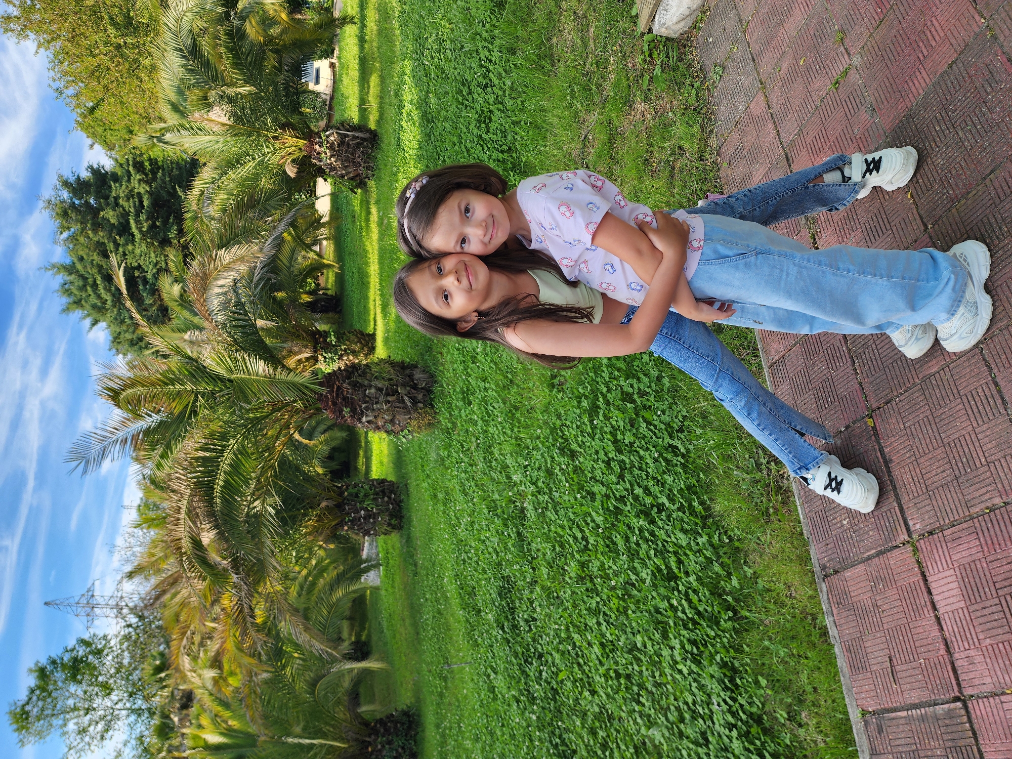 The image features two young girls embracing each other in an outdoor setting in Sochi, Russia. The girl on the left, with long dark hair, wears a light green tank top and blue jeans, smiling gently. She has her arms wrapped around the other girl. The girl on the right, with shorter dark hair and a pink headband, wears a patterned light-colored t-shirt and blue jeans, resting her head on the first girl's shoulder and smiling broadly. Both girls are wearing white sneakers.
They are standing on a red brick pathway next to a vibrant green lawn or field of clover. The background is lush with vegetation, dominated by numerous tall palm trees and other green trees, including some coniferous varieties, typical of a subtropical region. Above, the sky is clear and blue with scattered wispy white clouds. In the distance, an electricity pylon is visible against the sky. The bright natural lighting indicates it is daytime, likely a sunny late morning or afternoon. The girls appear happy and affectionate, enjoying their time together outdoors. There is no visible text in the image.