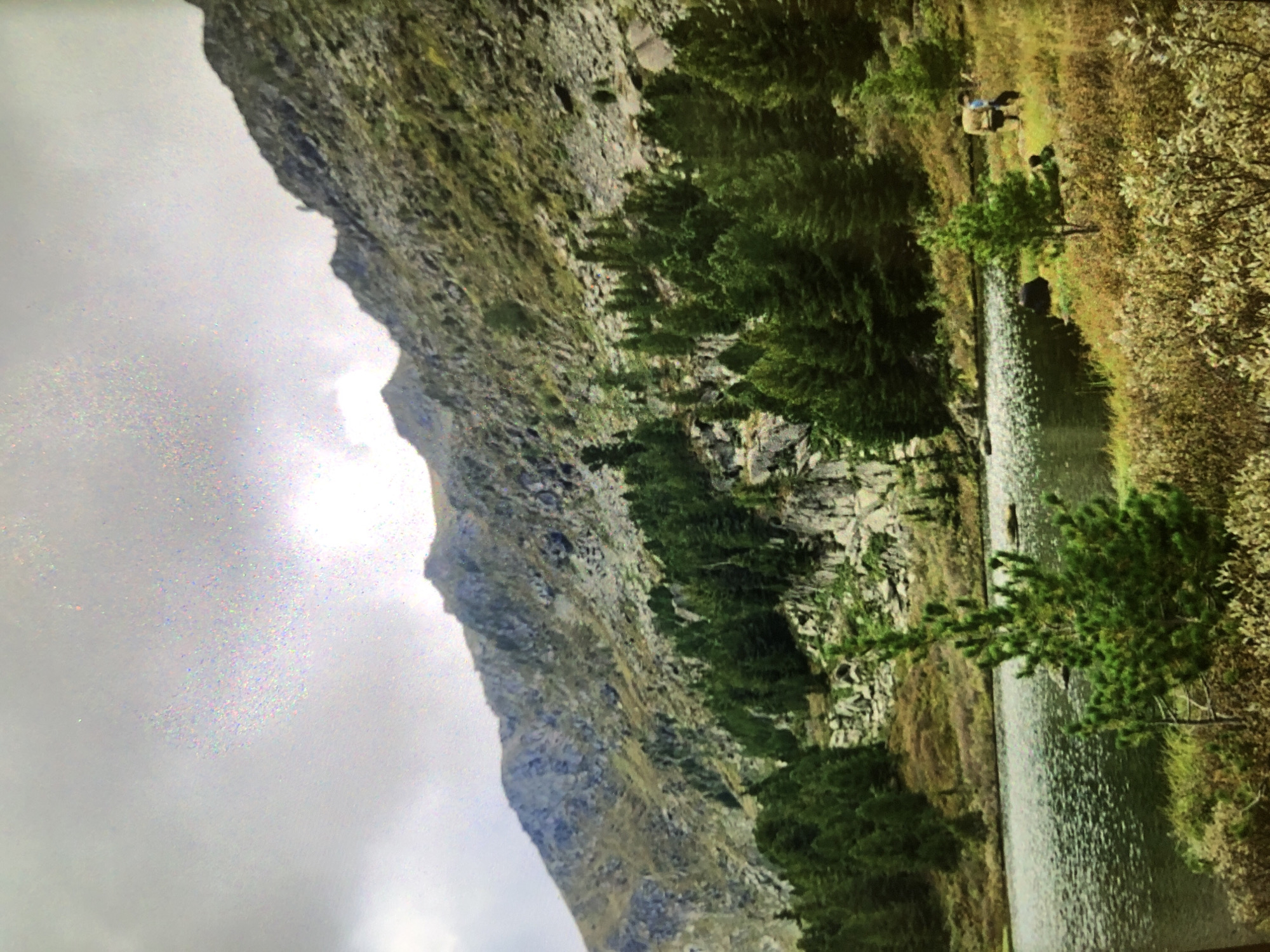 The image presents a mountainous landscape adjacent to a body of water. A steep, rocky mountain face dominates the left side of the frame, transitioning into dense vegetation and trees along the water's edge. The water surface reflects ambient light. On the opposite bank, there are areas of tall grass and shrubs. In the background, there are human figures standing near the water. The sky is overcast. The location appears to be a remote natural environment, possibly in a mountainous region.