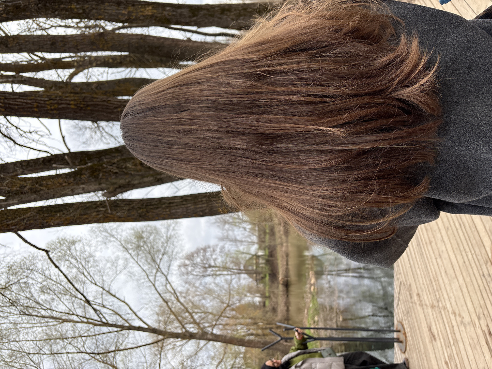 The image depicts an outdoor scene in Sdt Shcherbakovka, Russia, showing a person from behind, with long, wavy brown hair and wearing a dark grey coat. The individual is standing on a light-colored wooden deck, looking out towards a body of water, likely a river or lake. The background features numerous deciduous trees with bare branches against an overcast sky, suggesting a setting in late winter or early spring. Along the distant banks of the water, more trees are visible, contributing to a natural, somewhat muted landscape. In the lower right portion of the frame, another person is partially visible, appearing to be sitting or tending to equipment that resembles fishing rods or tripods, indicating an outdoor leisure activity such as fishing. The water surface is relatively calm, reflecting the muted tones of the sky and distant banks. The overall atmosphere is serene and focuses on nature and outdoor recreation. No text is visible in the image.