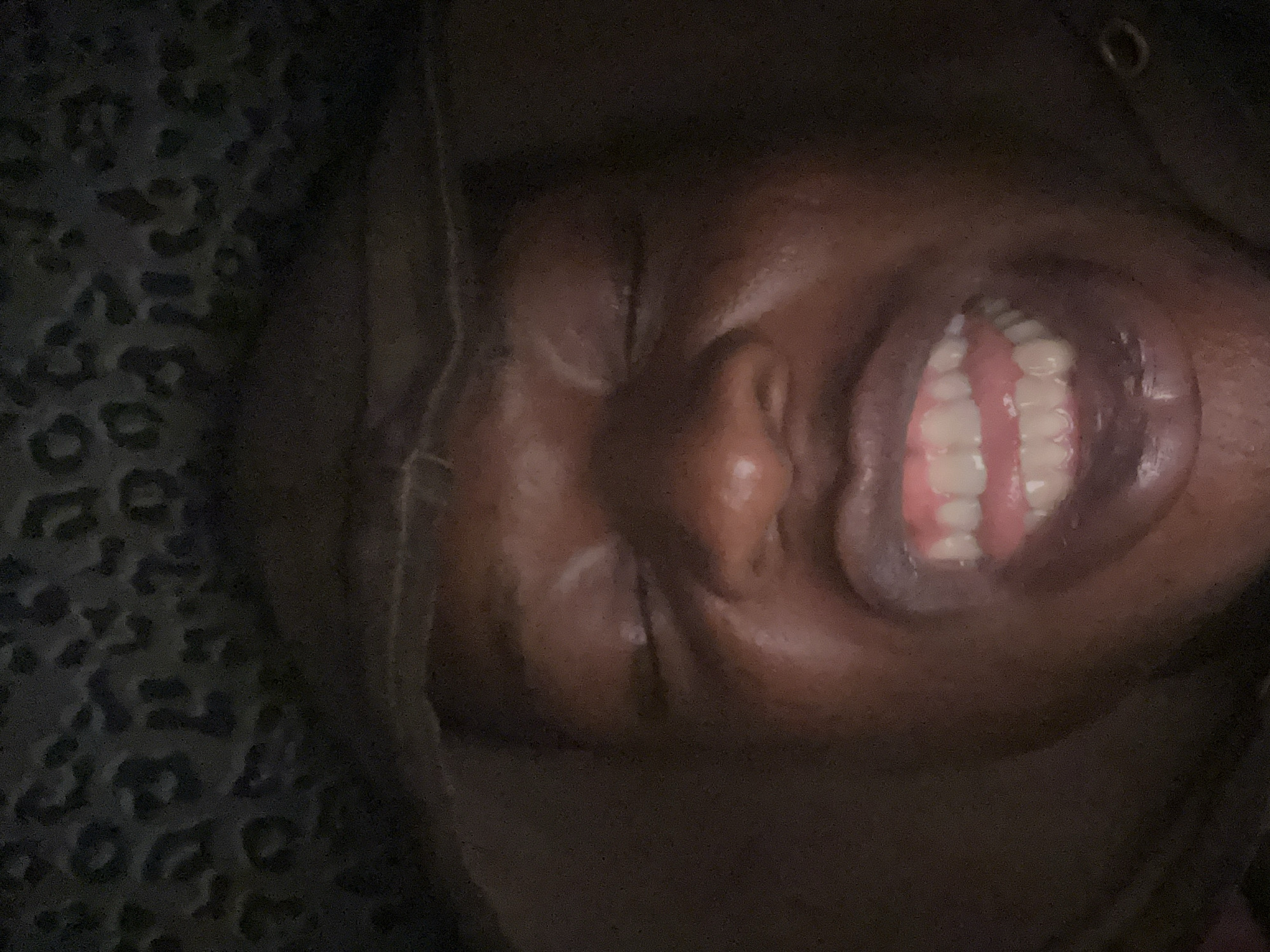 The image is a close-up, low-angle shot of a person's face, taken indoors. The subject has dark skin and their eyes are closed. Their mouth is open, revealing teeth, and their lips appear glossy. They are wearing a hooded garment, with the hood pulled up around their head, casting shadows on their face. The background is dark and appears to be a patterned fabric, possibly a blanket or upholstery, with a leopard-like print visible in the upper portion of the image. The lighting is dim, creating a somber mood. The scene appears to be a personal, candid moment, without any obvious indication of a specific location within Ikorodu, Nigeria, beyond being an indoor setting.