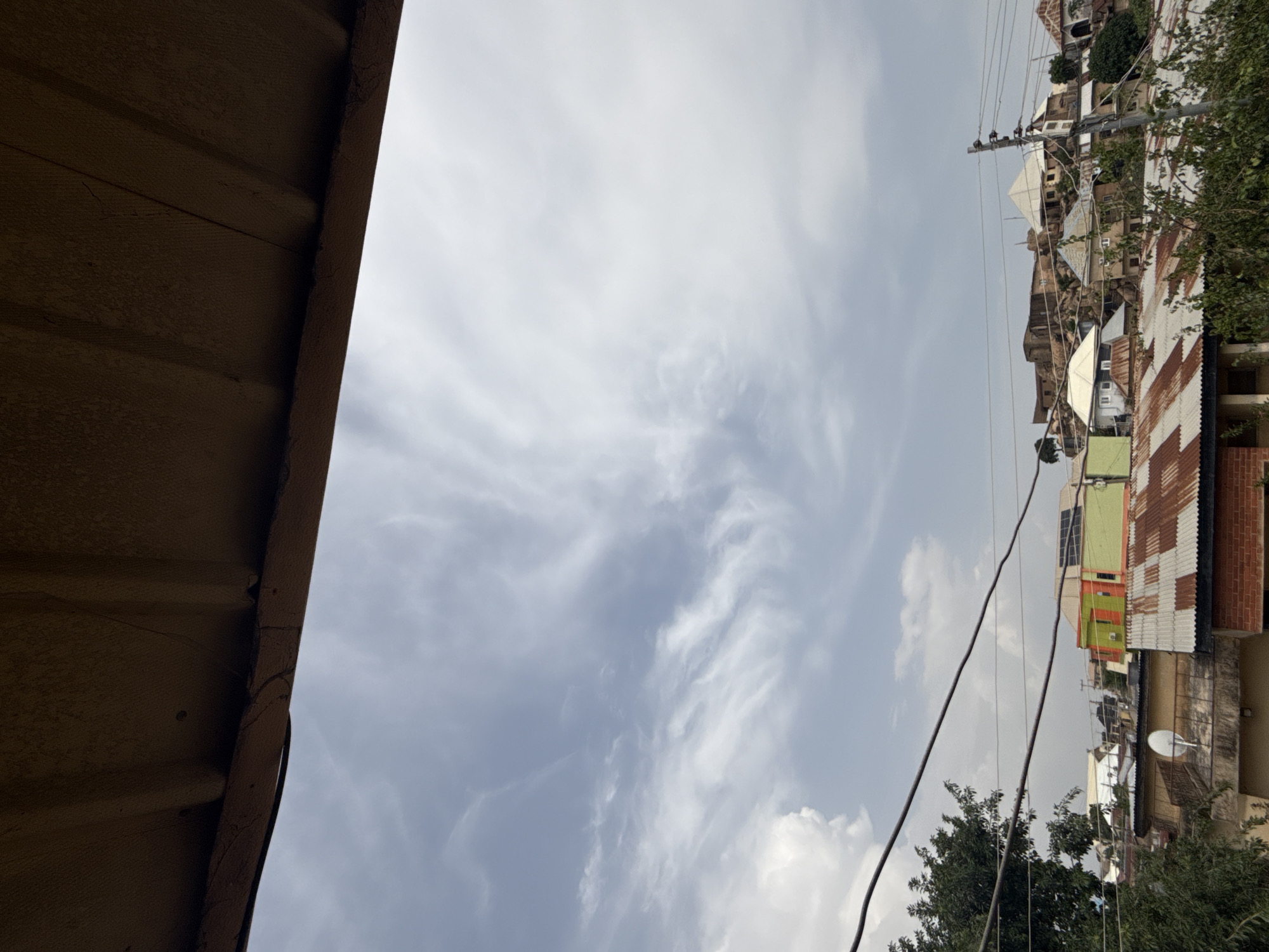 The image depicts an overhead view of a densely populated urban area. In the foreground, a corrugated metal roof overhangs the scene, casting a shadow. Below this, a network of power lines stretches across the frame, obscuring a portion of the sky and the cityscape. The sky itself is partly cloudy, with wispy cirrus clouds scattered across a pale blue backdrop.
The middle ground of the image showcases a collection of buildings with varied roofing materials, including corrugated metal and what appears to be tile. Some structures are brightly colored, with orange, green, and yellow accents, while others are more muted in tone. Satellite dishes are visible on some rooftops. Trees with dense foliage are interspersed among the buildings, adding a touch of greenery to the urban landscape.
The background features a hilly terrain with more structures built into the slopes. The architecture in the background suggests a mix of traditional and modern construction. The overall impression is one of a bustling, lived-in city with a strong sense of community evident in the close proximity of the buildings. The image captures a moment of a typical day in this urban environment.
