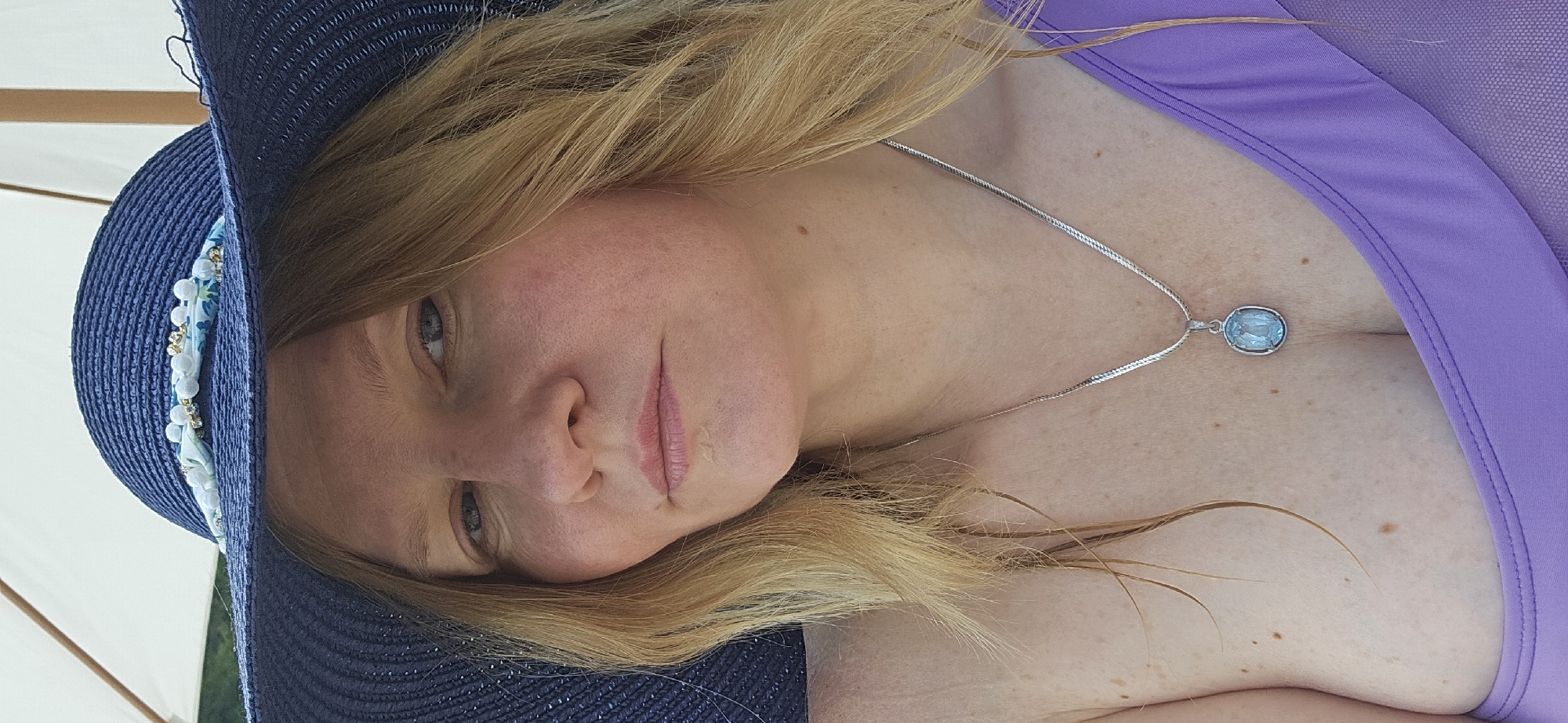 A woman with blonde hair and blue eyes is pictured wearing a blue straw hat and a purple swimsuit. She is also wearing a silver necklace with a light blue pendant. The woman appears to be outdoors, possibly at a beach or poolside, with a white umbrella visible in the background. The setting suggests a warm, sunny day. The image was taken in Novomikhaylovsky, Russia.