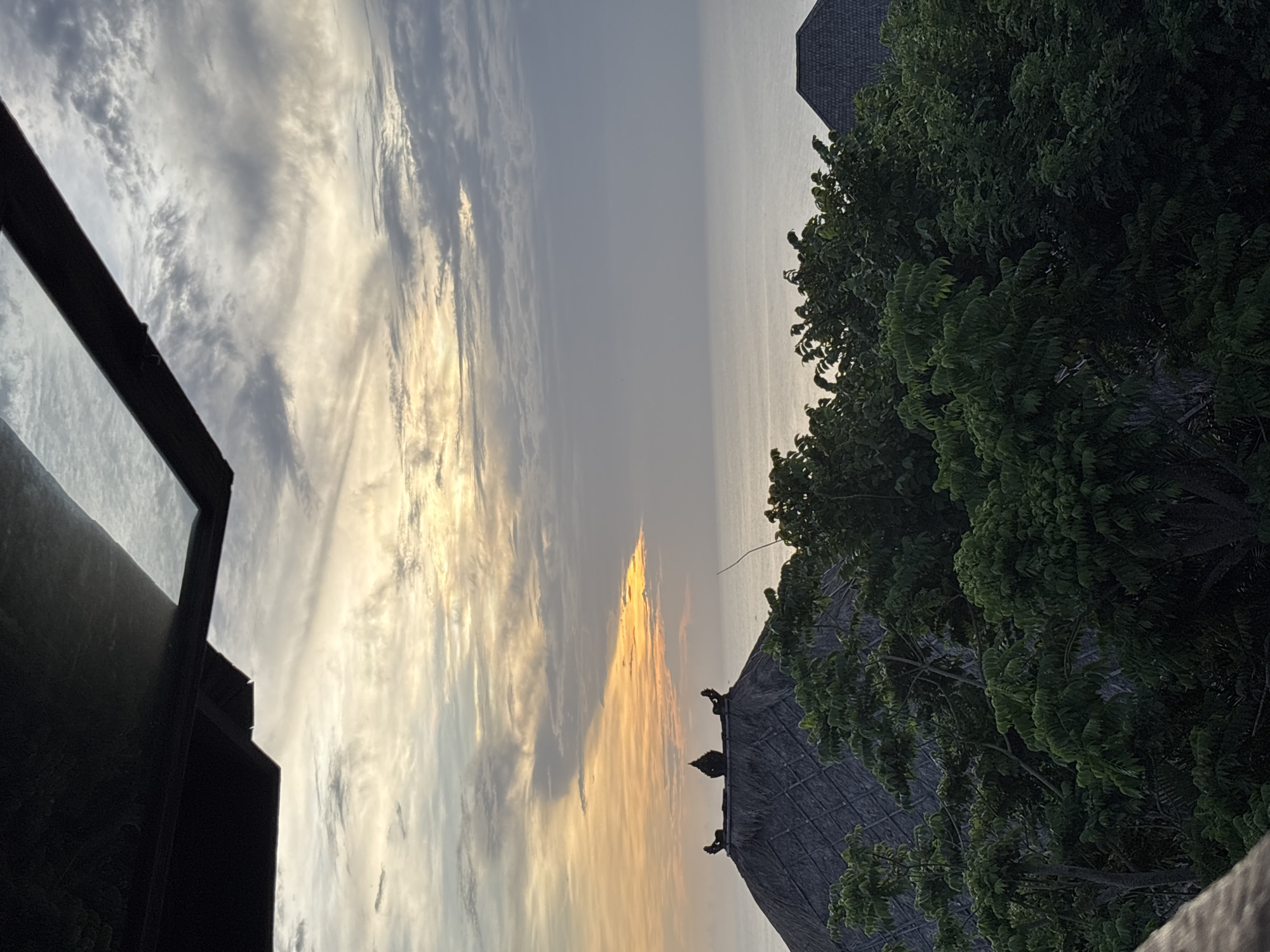 The image, taken in Kuta Selatan, Indonesia, captures a view of the sky at sunset. The sky is filled with streaks of yellow and grey clouds, with the sun appearing to shine through the clouds in the middle. In the foreground, on the right side, there are green trees and a roof with decorative ornaments. On the left side, a dark structure is visible, possibly a building or part of a building. The horizon line is faintly visible in the background, indicating the ocean.