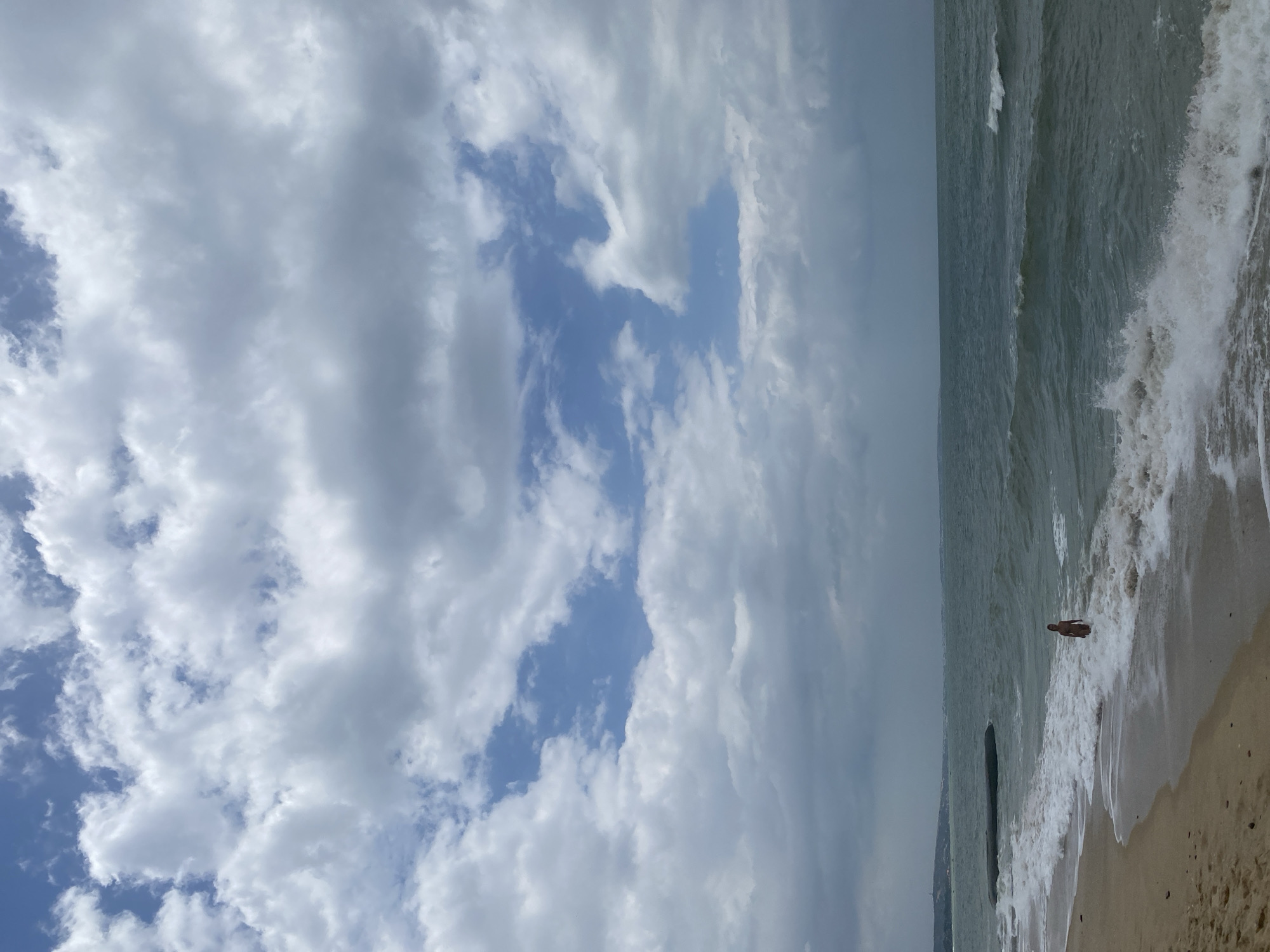 The image depicts a beach scene in Mũi Né, Vietnam. A person, appearing to be a child, stands in the shallow water near the shoreline, facing the ocean. The water is moderately wavy with white foam washing ashore. The beach itself is sandy with some small pebbles visible. In the distance, a dark, cylindrical object protrudes from the water, and a coastline with hills can be seen. The sky is filled with a mix of white and gray clouds, with patches of blue sky showing through. The overall atmosphere is overcast, suggesting a potentially cloudy or stormy day.