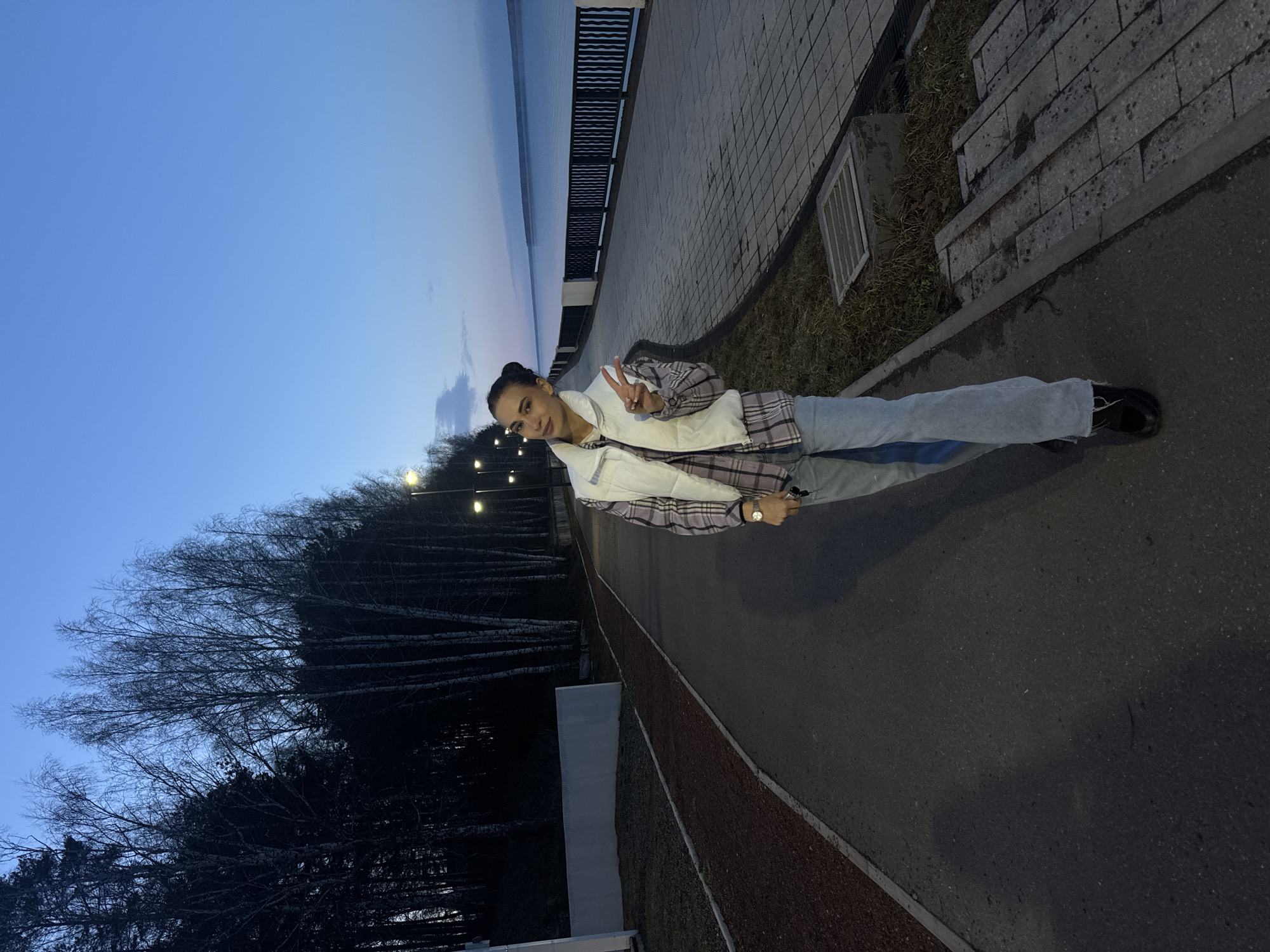 A young woman stands on a paved outdoor pathway next to a large body of water in Chaikovsky, Russia, during twilight or early evening. She is facing the camera, smiling faintly, and making a peace sign with her right hand. Her attire consists of a plaid shirt, a white puffer vest, light blue jeans, and dark shoes. Her dark hair is pulled back, and a watch is visible on her left wrist.
To the woman's left, a dark metal fence runs along the edge of the pathway, separating it from the wide, calm expanse of water. The water's surface reflects the deep blue and subtle orange tones of the twilight sky. To her right, the pathway is bordered by a strip of reddish gravel or mulch, beyond which lies a grassy area featuring a mix of bare deciduous trees, possibly birches, and darker evergreen trees. Several artificial lights, appearing as streetlights or decorative path lights, glow in the distance among the trees and along the waterfront.
The sky is a clear, deep blue, transitioning from day to night, with a hint of lighter tones near the horizon over the water. The ground is a dark asphalt pathway. To the far right, near the grass, there are some concrete elements, including a grate, possibly part of a drainage system. The scene suggests a calm evening in a park or promenade area. No visible text is present in the image.