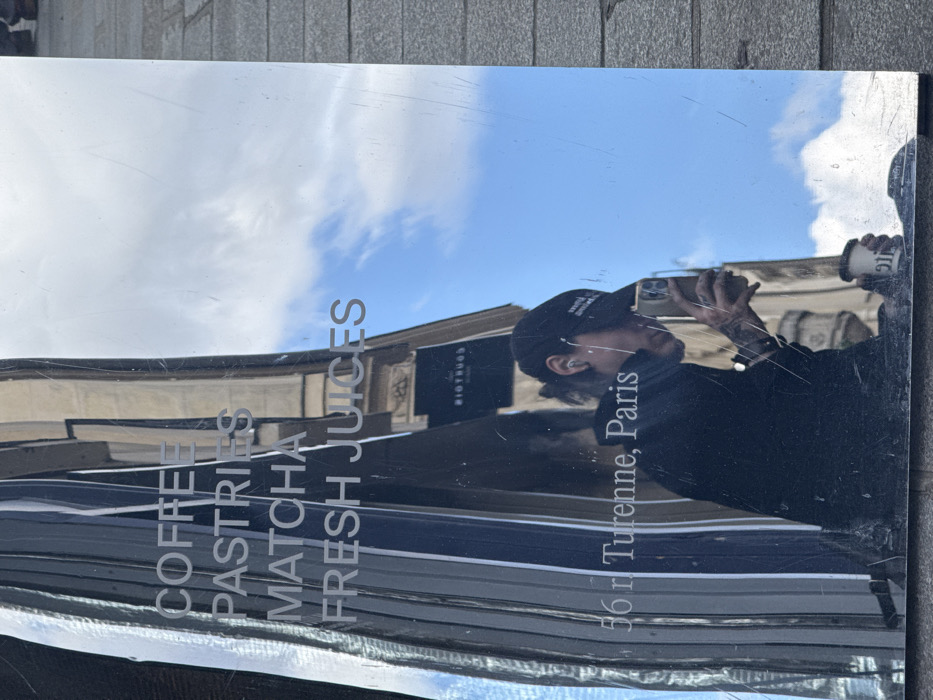 The image displays a distorted reflection on a vertical, highly polished surface, likely metal or mirrored glass, in an outdoor urban setting. Engraved or affixed to this surface are the words "COFFEE," "PASTRIES," "MATCHA," and "FRESH JUICES," along with the address "56 r Turenne, Paris."
Reflected in the surface is an individual, seen in profile, wearing a dark baseball cap with white lettering, dark long-sleeved attire, and white in-ear headphones. The person holds a light-colored smartphone in their right hand, angled as if capturing an image or video, and a white disposable coffee cup with "911" branding visible in their left hand. Tattoos are present on the individual's left forearm.
The reflected background includes a partially cloudy blue sky and sections of traditional Parisian building architecture, featuring windows and a dark sign with "RIOTTUGG" in white text. A light-colored paved ground surface is visible to the right and at the bottom of the reflective panel. The confirmed location is 56 Rue de Turenne, Paris, France.