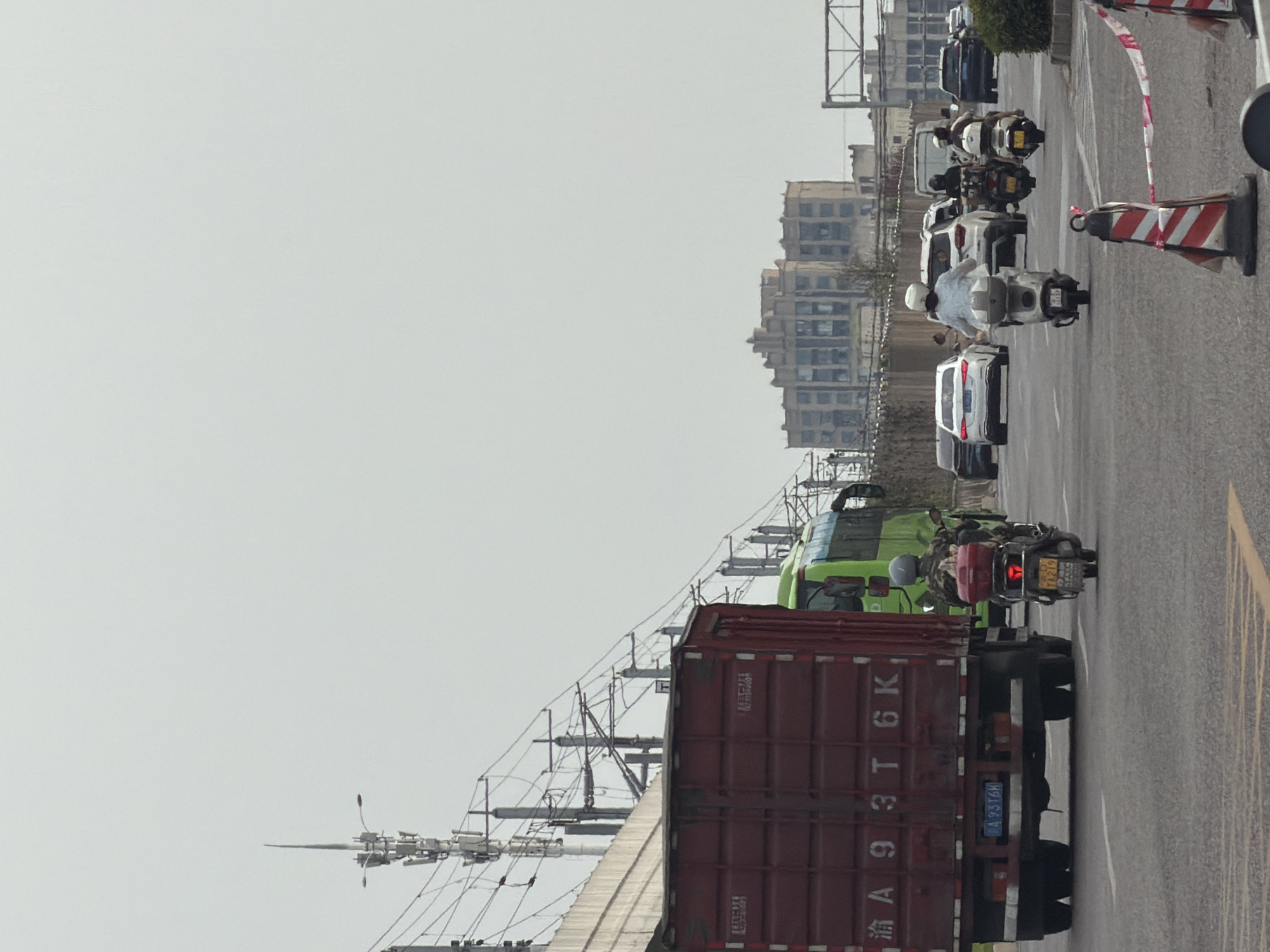 The image captures a street scene in Jiangjin, China, under an overcast sky. In the foreground, a large red shipping container truck is visible, with its license plate reading "A 93T6K." To the right of the truck, a motorcycle with a rider wearing a camouflage jacket and a helmet is seen. Behind the truck, a green bus is partially visible. Further down the road, several cars and motorcycles are in motion. On the left side of the image, a tall communication tower stands prominently, surrounded by power lines. In the background, a multi-story building with a distinct architectural style is silhouetted against the sky. Construction barriers with red and white tape are present in the lower right portion of the frame, suggesting ongoing work. The overall impression is of a typical urban traffic environment.