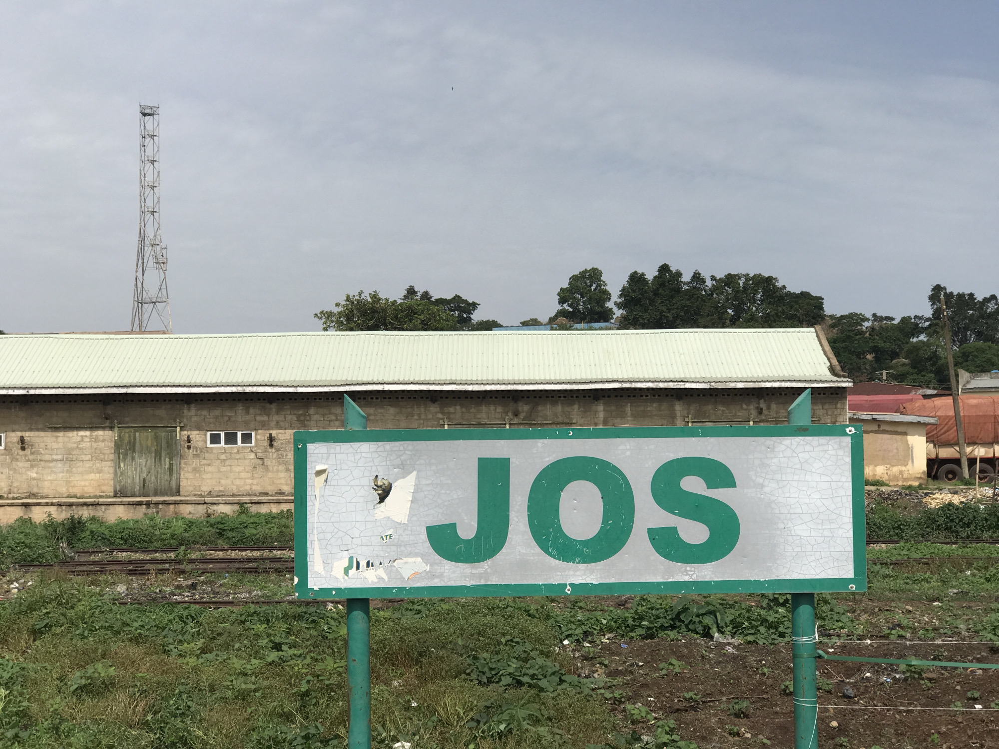 This is a forensic, matter-of-fact description of the image, which appears to be taken in Jos, Nigeria.
The image depicts a sign with the word "JOS" in large green lettering against a distressed silver background, framed by a green border. The sign is supported by a green metal post. To the left of the sign, a long, light-colored building with a corrugated metal roof is visible. This building has a large, weathered wooden door and several small, rectangular windows. The building appears to be constructed of brick or stone blocks.
In front of the building and extending towards the foreground, there are several sets of railway tracks, suggesting a railway station or yard. The area around the tracks and in the foreground is covered in overgrown green vegetation and some patches of disturbed soil. In the background, beyond the building, a tall metal tower structure is visible against a cloudy sky, along with dense foliage from trees. Further to the right, parts of other buildings and what appears to be a large truck with a red tarp can be seen. The overall environment suggests an outdoor scene in a somewhat industrial or semi-rural setting within Jos. There are no people visible in the image.
