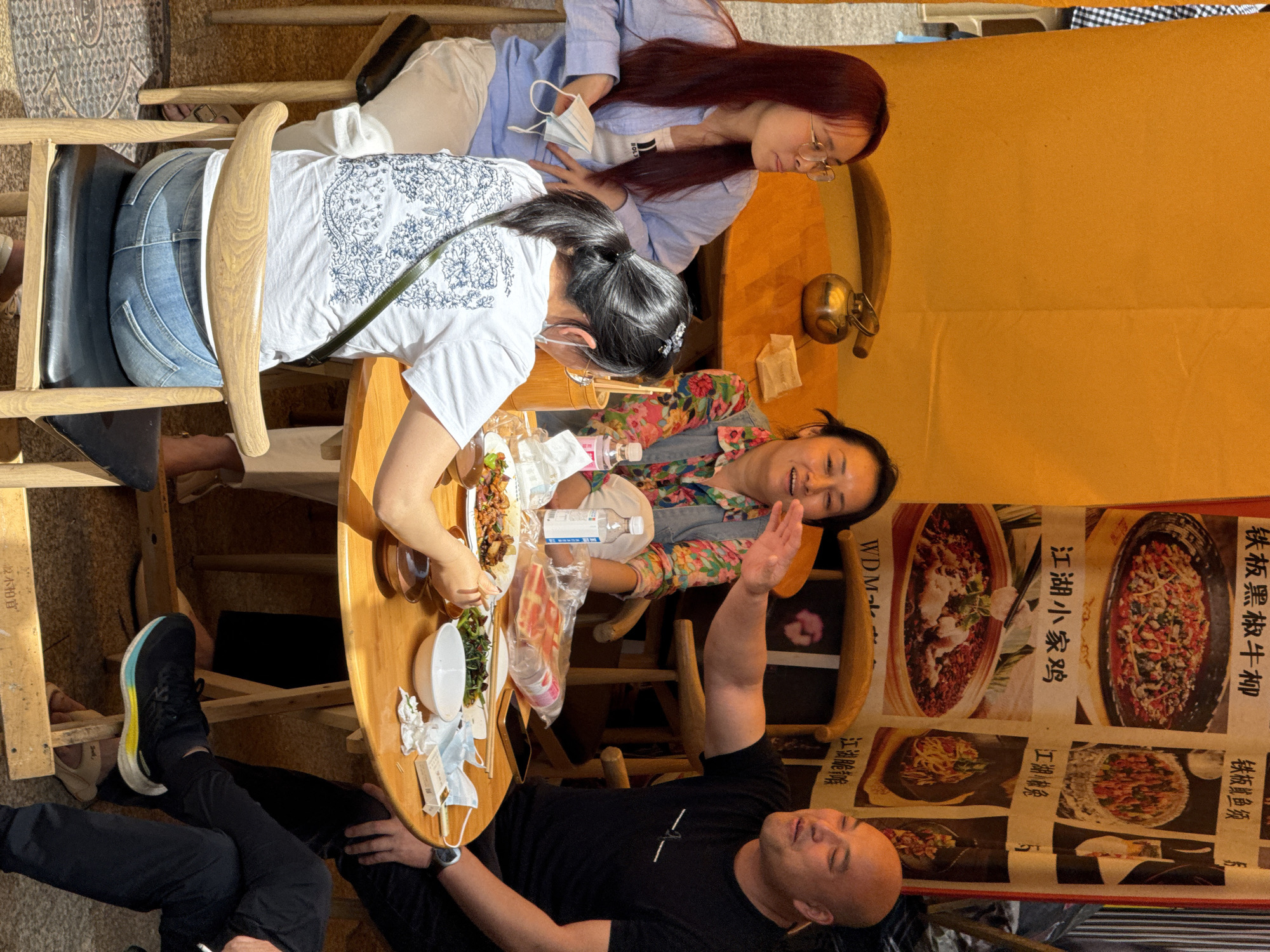 Here is a forensic description of the image:
**Scene:** An indoor shot appears to capture a group of people dining in a restaurant. A poster with food images and Chinese characters is visible on a wall.
**People:** Four individuals are depicted. Two women are seated at the table. One has long red hair and wears glasses, while the other has black hair tied back. A third woman is partially visible, smiling. A man with a shaved head is lying on the floor, gesturing with his right arm.
**Objects:** The table is laden with food, including dishes of cooked items and a white bowl, potentially filled with vegetables or salad. A transparent plastic bag, possibly containing takeout, sits on the table. Other objects include a water bottle, chopsticks, what appears to be a package of cigarettes, and multiple disposable face masks.
**Actions/Interactions:** The red-haired woman appears to be looking down, possibly at her food or phone. The woman with the black hair is actively eating with chopsticks. The smiling woman is seemingly engaging with the man on the floor, who is raising his arm.
**Environment:** The restaurant has wooden tables and chairs.
**Location:** Given the Chinese characters on the poster, and OCR output indicating "江油脆喉" which translates to "Jiangyou Crispy Throat", the location is identified as being in the city of Yuzhong District, China.