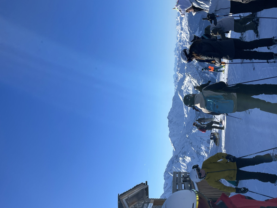 The image captures a vibrant winter scene in the snowy mountains of Séez, France, under a clear, bright blue sky. The shot is composed from a slightly elevated perspective, looking out over vast snow-covered peaks that stretch across the horizon.
Several people, presumably skiers, are gathered in the foreground and mid-ground of the image, equipped with ski gear including helmets, goggles, skis, and poles. In the immediate foreground-right, a person in a bright yellow jacket and black pants stands on skis, wearing a white helmet. To their left, another person in a white helmet and red jacket is partially visible. Further back, a skier with a light blue backpack and a black helmet stands, while another individual wears a dark jacket and a white helmet. Towards the top-right, a person stands out with a distinctive white, fluffy unicorn-themed helmet cover. All individuals appear to be taking a break or waiting on a ski slope, facing the expansive mountain range.
The weather is bright and sunny, with strong sunlight casting clear shadows. Visible sun rays pierce through the upper left portion of the sky, indicating a strong light source. The only clouds are very faint, high-altitude wisps.
In the foreground-left, a rustic wooden structure, possibly a signpost or part of a small hut, is visible. Text on this structure includes "EN COURS" (in progress) and "PISTE VERTE" (green slope), suggesting they are on an easy ski run. Another wooden barrier or fence is also present near the foreground skiers. The overall atmosphere conveys a cheerful day for winter sports amidst a breathtaking alpine landscape.