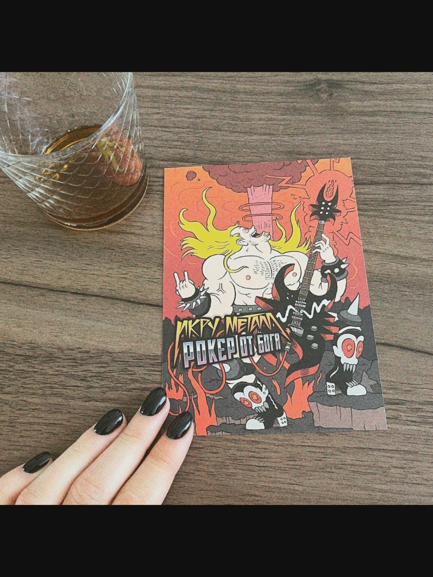 A close-up, overhead shot shows a person's left hand with glossy black nail polish reaching towards a postcard placed on a wooden surface. The postcard features a cartoon illustration of a muscular, blonde-haired character with a guitar, set against a backdrop of flames and smoke. The text on the postcard appears to be in Russian. To the left of the postcard, a glass containing a dark liquid and what looks like strawberries sits on the wooden surface. The lighting is warm and natural. The specific location in Saint-Août, France, is not discernible from the image.