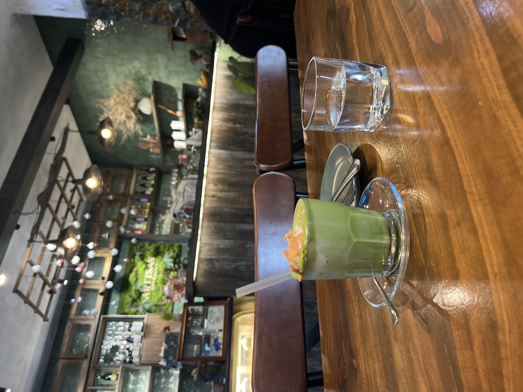 A wooden table in a restaurant setting features a glass of water and a green drink with a straw and what appears to be foam or toppings. The drink sits on a saucer with a spoon. The table is adjacent to chairs and a bar area. The restaurant has visible shelves with items, lighting fixtures, and greenery. A sign with the word "ARTISAN" can be seen. The location is Nha Trang, Vietnam.