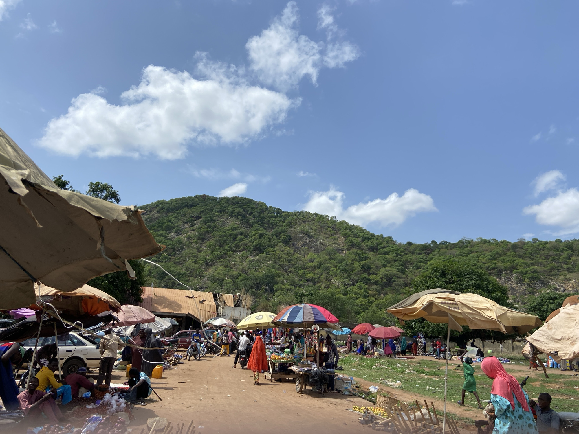 This image depicts a bustling open-air market scene in Mararaba, Nigeria, set at the base of a large, verdant, forested hill under a bright blue sky with scattered white clouds, indicating daytime. The ground is unpaved and appears dusty.
Numerous people are present, including vendors and shoppers of all ages. Many are dressed in casual attire, with some individuals wearing traditional West African clothing such as long robes and hijabs. People are engaged in various market activities, including selling goods, walking through the market, and sitting on the ground tending to their merchandise.
The market is characterized by a multitude of colorful umbrellas—beige, red, blue, and striped—providing shade for the stalls and people. Under these umbrellas, a variety of goods are displayed, including large piles of reddish onions, yellow fruits (possibly mangoes or lemons), and other packaged items on carts and makeshift tables.
Vehicles such as cars and motorcycles are visible in the background, suggesting ease of access to the market. In the mid-ground, a person in a bright orange garment is walking through the crowd. On the far left, individuals are gathered around goods, with a white car parked nearby. Towards the right, a person in a pink hijab and blue patterned dress is visible, with children nearby.
Some structures with corrugated iron roofs are seen at the base of the hill, and there is some visible debris and litter on the ground. Faintly visible on one of the large beige umbrellas in the middle-right of the frame is the partial word "HER". The scene conveys a vibrant atmosphere of commerce and daily life.